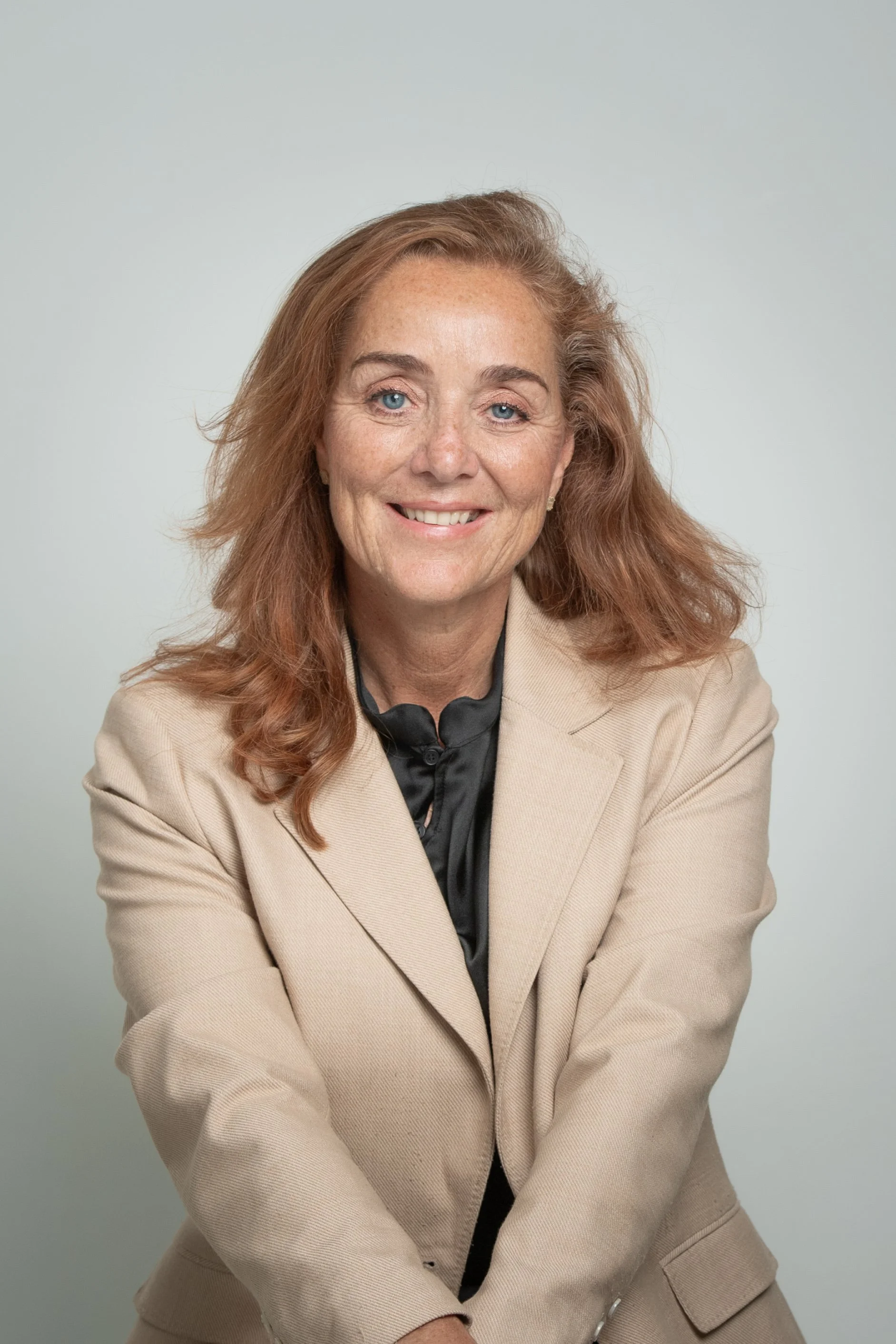 Een lachende vrouw met rood haar, gekleed in een beige jas en een zwarte blouse, poseert voor een lichtgrijze achtergrond.
