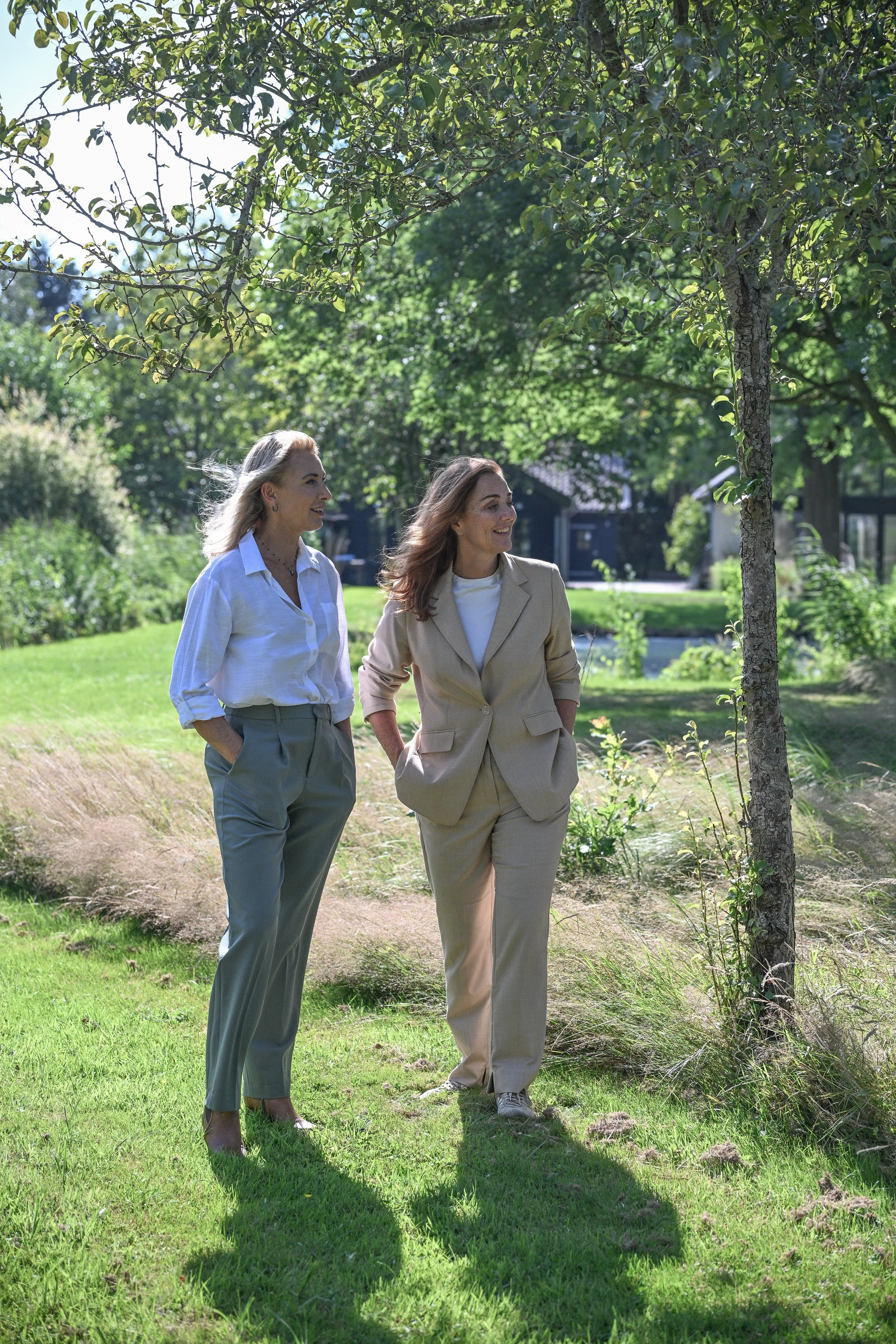 Twee vrouwen wandelen samen in een groene tuin op een zonnige dag, lachend en gekleed in casual zakelijke kleding.