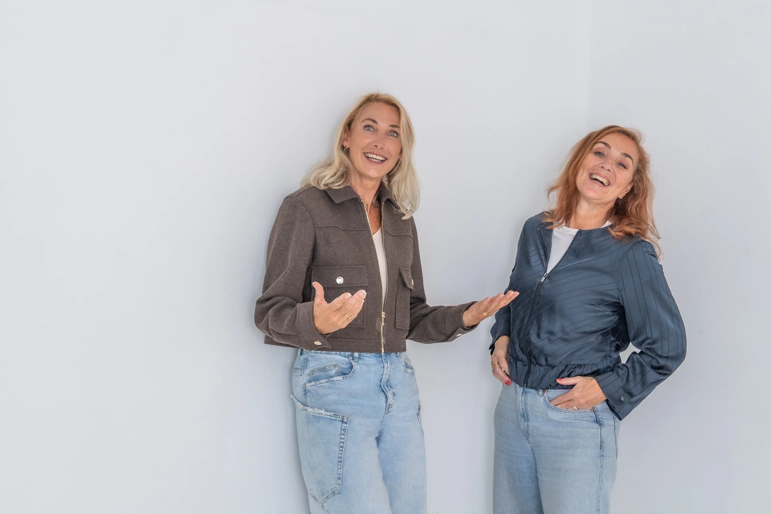 Twee vrouwen lachen en praten samen, gekleed in casual kleding, staand voor een witte muur.