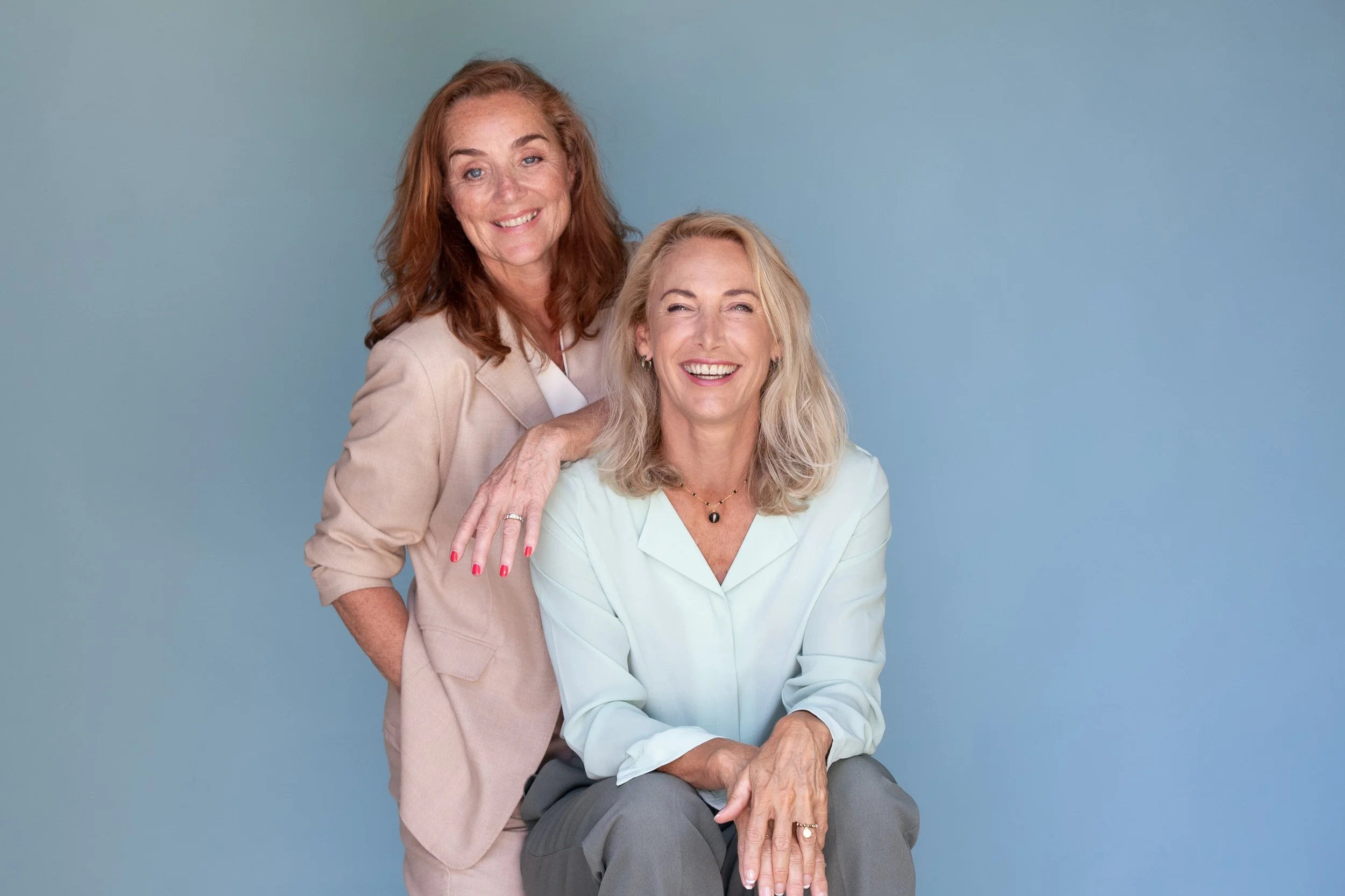 Twee vrouwen die lachen en poseren tegen een lichtblauwe achtergrond.