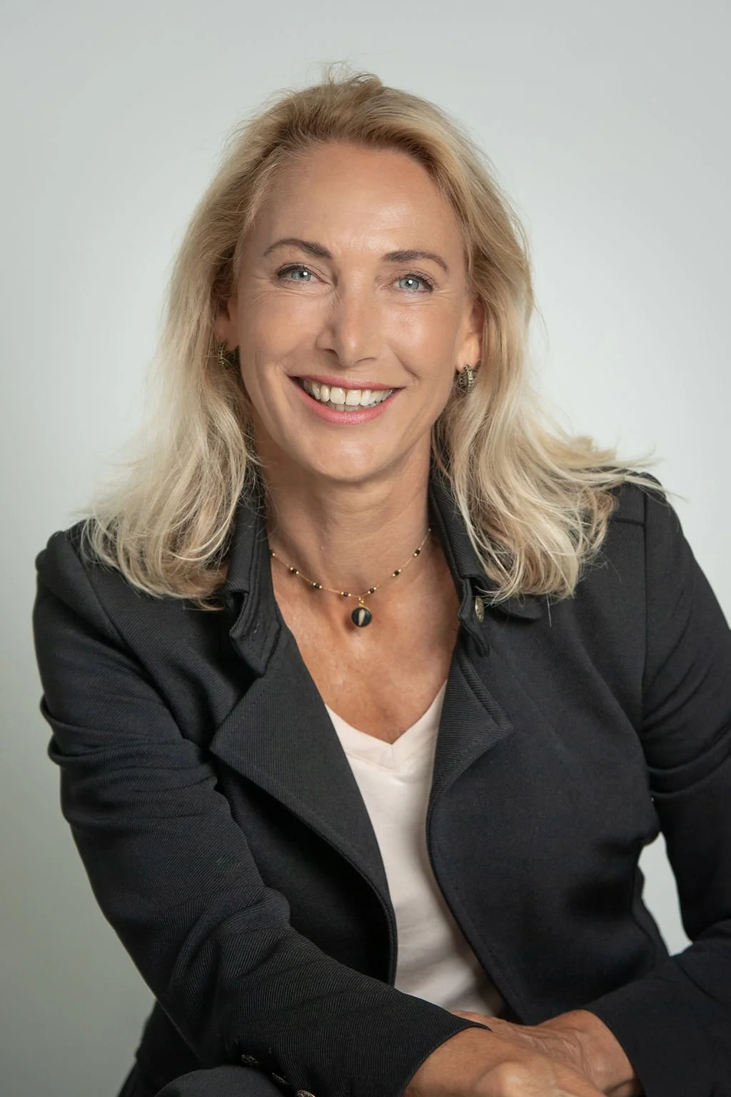Portret van een lachende vrouw met blonde haren, gekleed in een zwarte jas en witte top, tegen een lichtgrijze achtergrond.