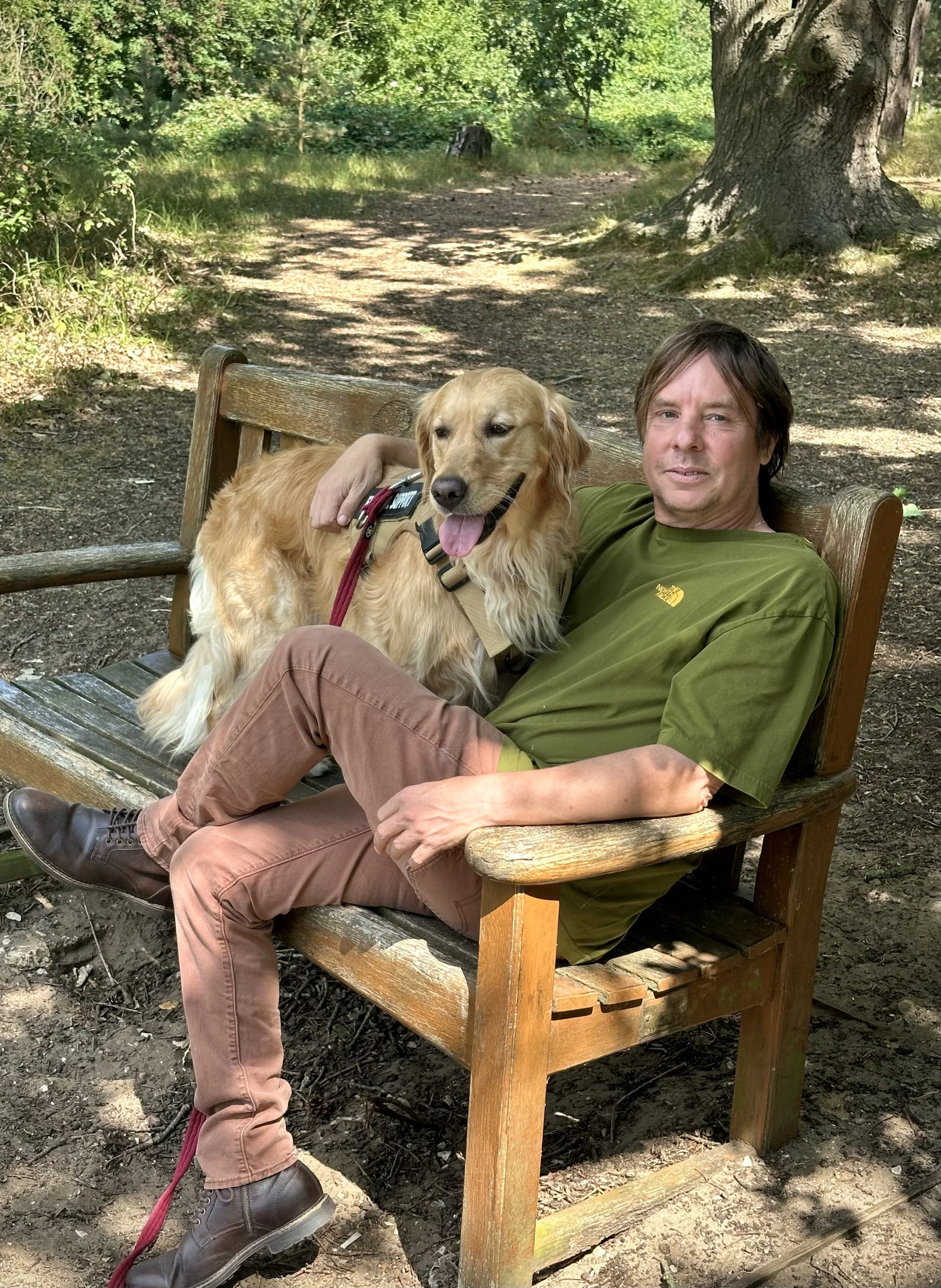 A man with dark brown hair wearing a green North Face shirt sitting on a wooden park bench with a golden retriever dog. The dog is standing on the bench with tongue out and a red leash, person’s arm is around the dog. The background is a lush green park with trees and sunlight filtering through leaves.