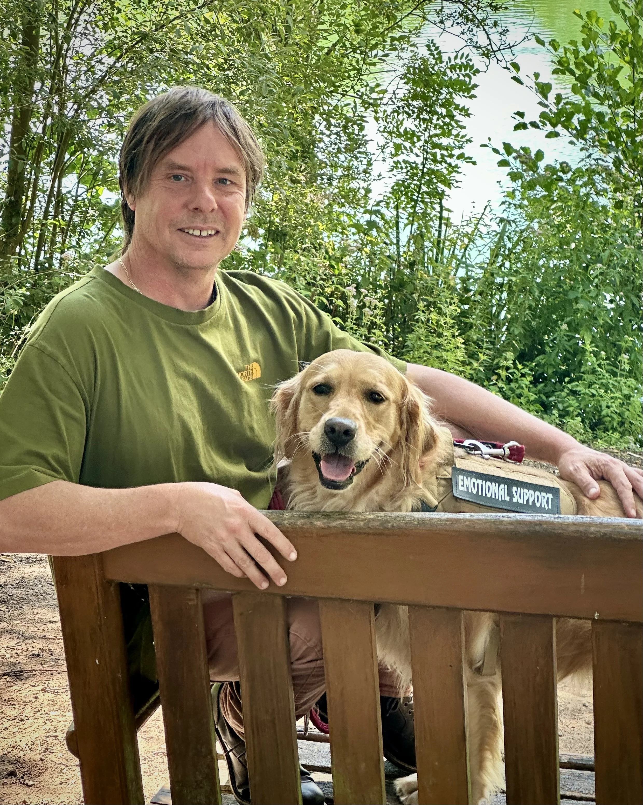 A man with brown hair wearing a green t-shirt sitting by a red and white retriever dog behind a wooden fence. There is greenery and water in the background.