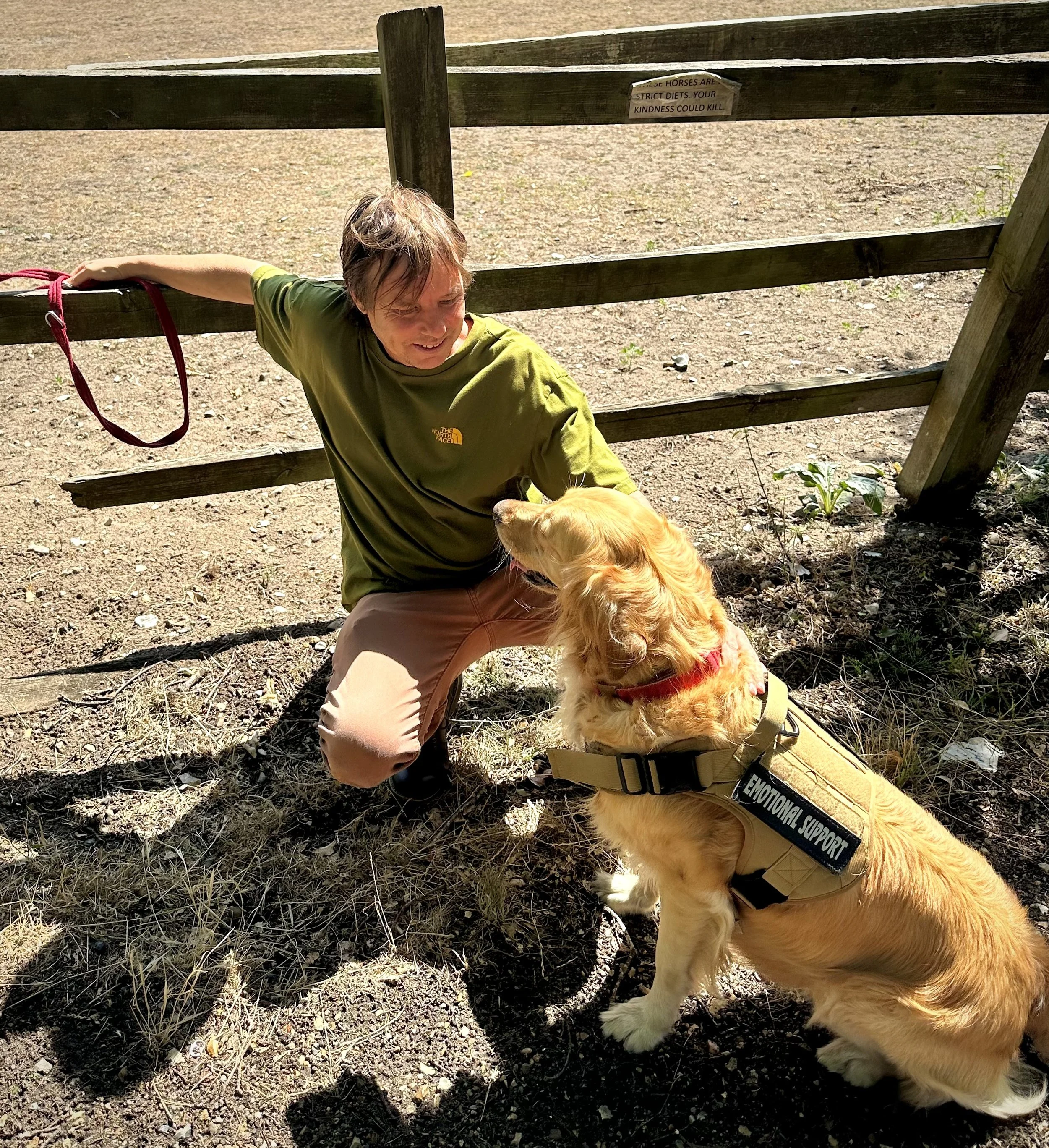 A person with short, light brown hair wearing a green North Face shirt and khaki pants is kneeling on a dirt ground next to a golden retriever dog with a red harness. The person is smiling and leaning towards the dog, which is wearing a vest labeled 'Emotional Support.' Behind them, a wooden fence with a sign and some small plants are visible, and the scene is outdoors in natural sunlight.