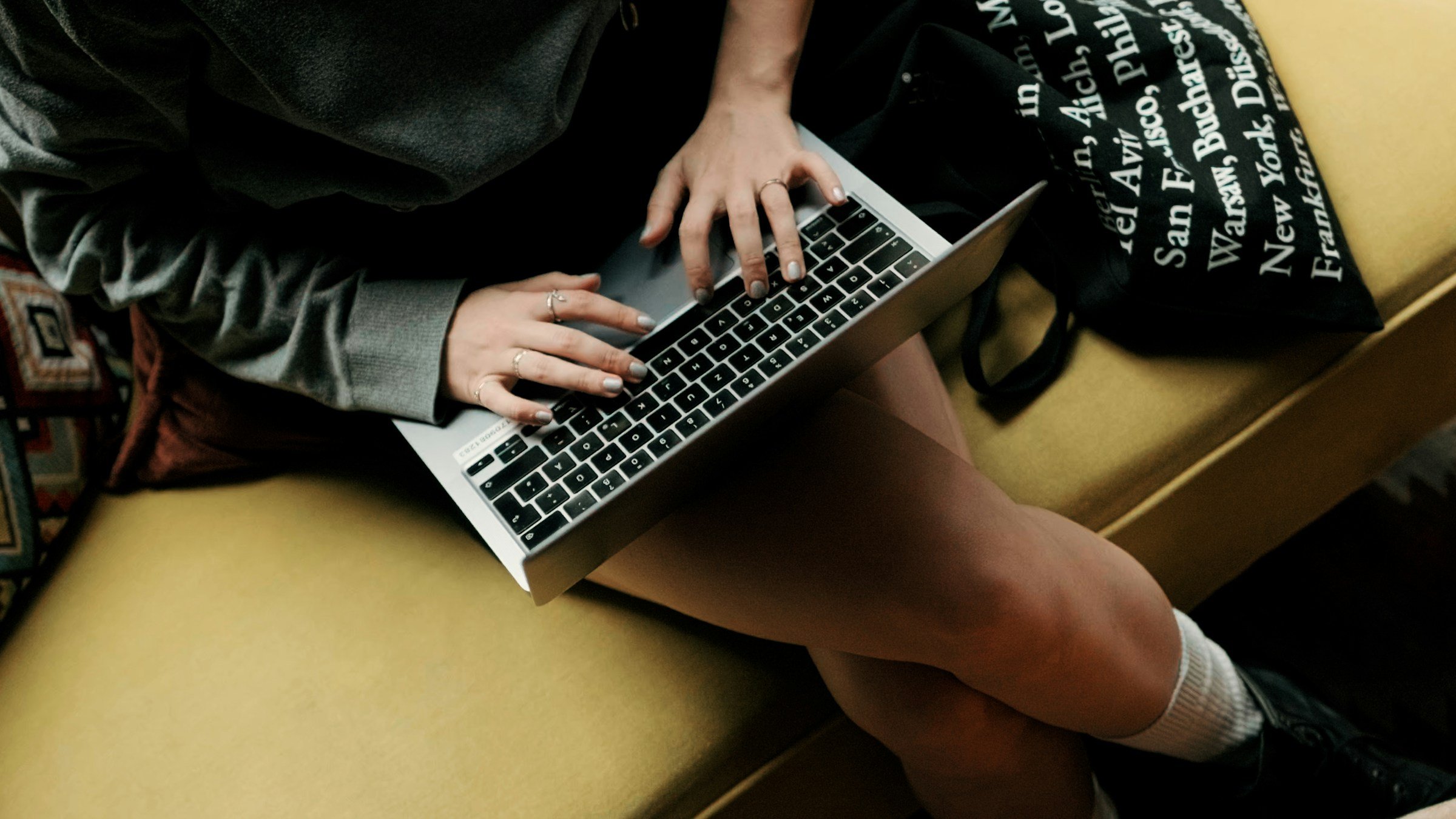 Person sitting cross-legged on a yellow couch, using a silver laptop on their lap, wearing a dark sweater and brown shorts.