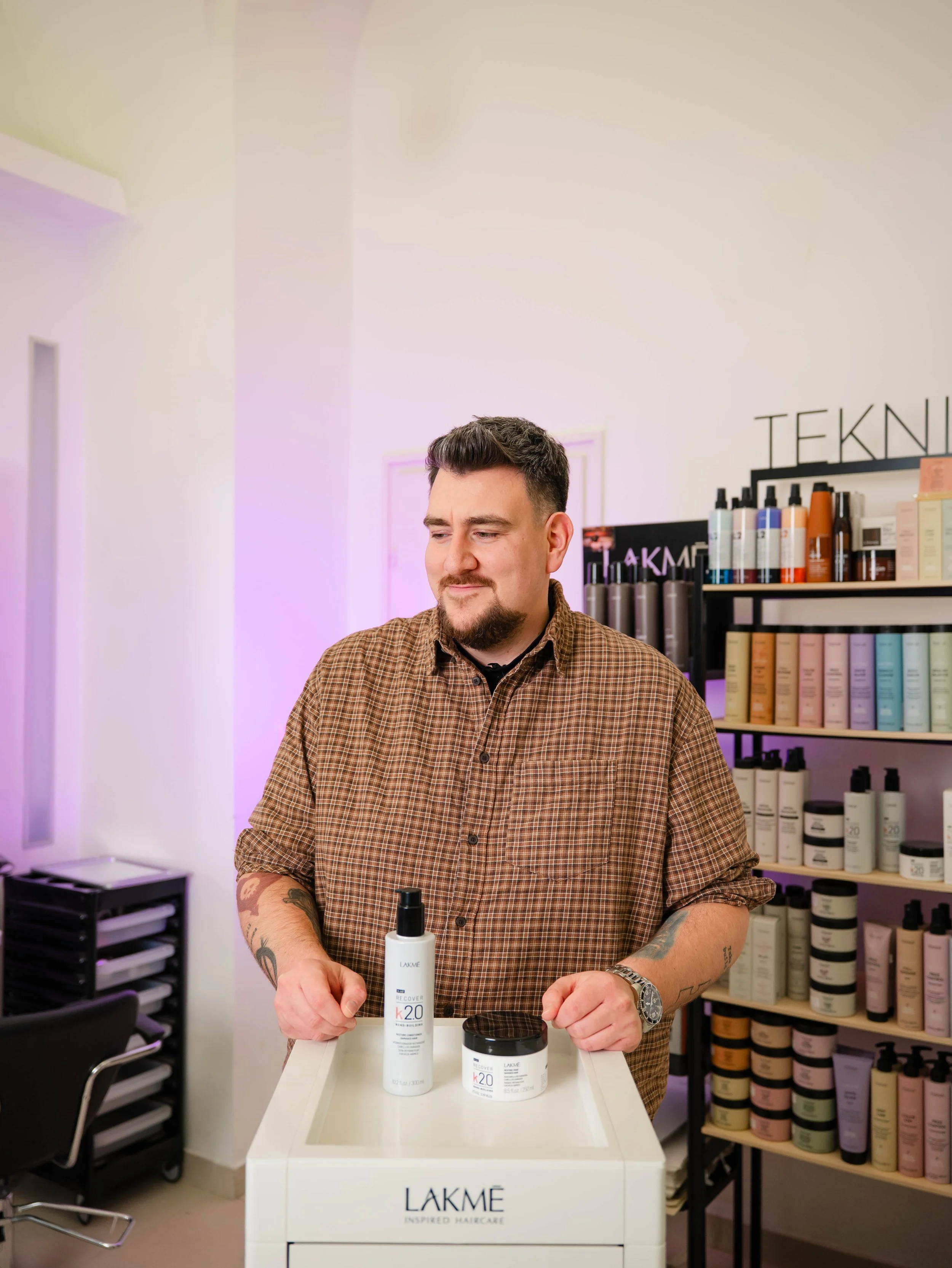 A man in a checkered brown shirt stands behind a table with skincare products, including a white bottle and a white jar, in a store with shelves of colorful hair and skincare products in the background.