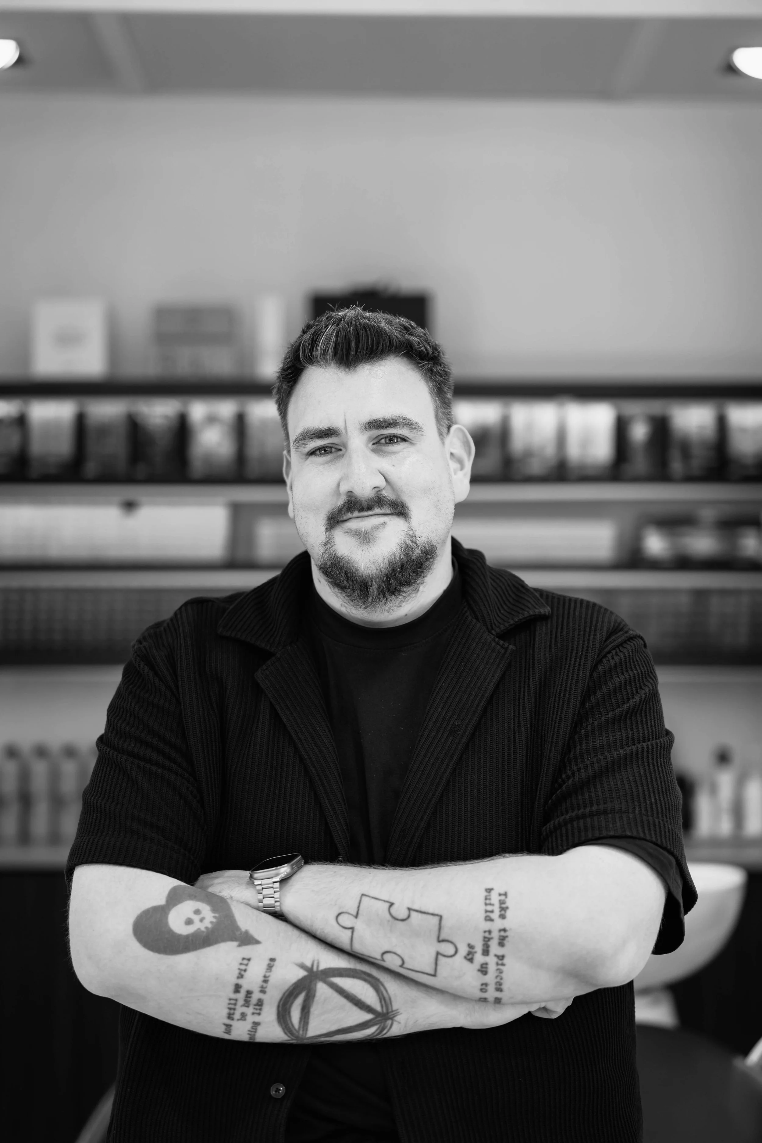 Black and white portrait of a man with short dark hair and a beard, sitting with arms crossed, in front of a background with shelves containing books and boxes, in an indoor setting.