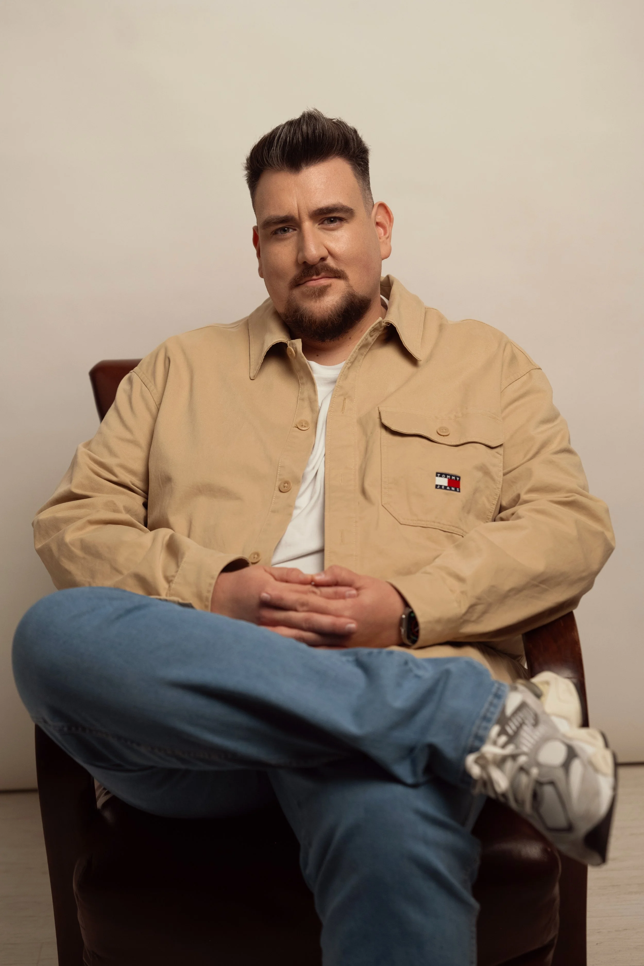 A man with dark hair, beard, and mustache sitting in a wooden chair against a plain light-colored wall. He is wearing a beige jacket with a Tommy Jeans logo, a white shirt, blue jeans, and white running shoes. He is sitting with his legs crossed and hands resting on his lap, looking directly at the camera with a neutral expression.