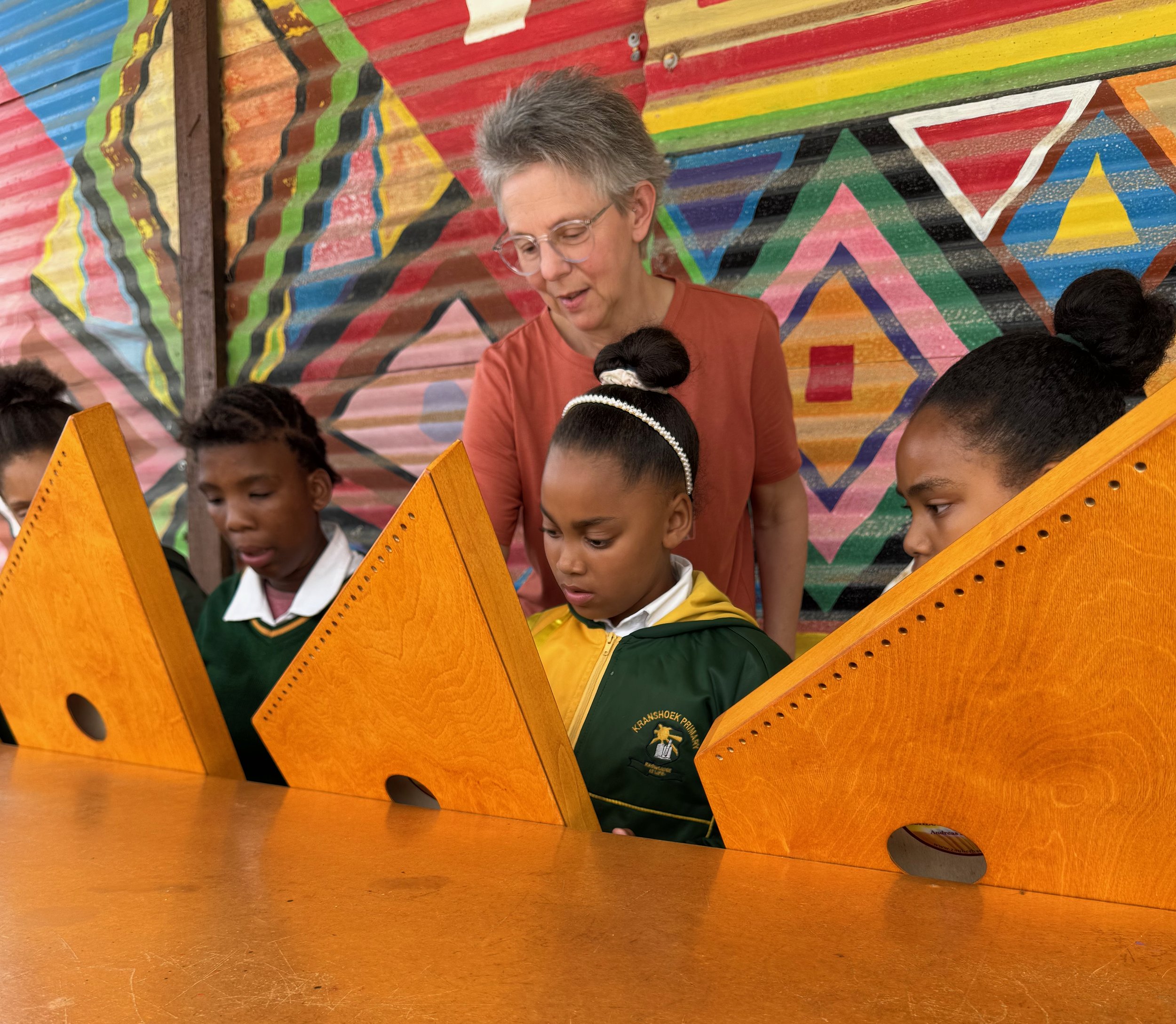 Eine Gruppe von Kindern sitzt an einem Tisch, während eine erwachsene Frau mit grau kurz geschorenem Haar und Brille sie anspricht, vor einem bunten, geometrischen Wandbild.