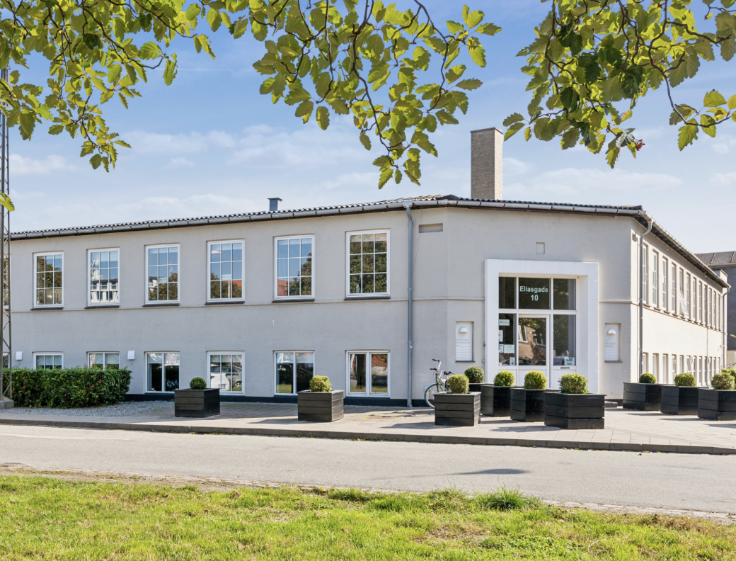 Hvidt tre-etagers kontorbygning med store vinduer, grønne planter i sorte plantekasser foran, en cykel parkeret ved indgangen, på Eliassgade 10, under en blå himmel med grønne træer i forgrunden.
