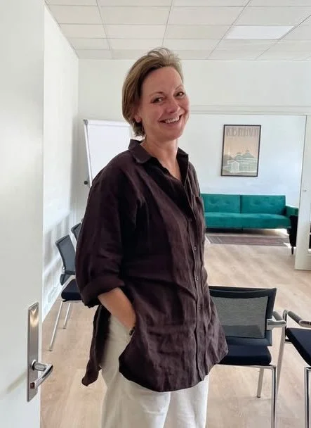 Kvindelig person står i et mødelokale med stole, en whiteboard og en grøn sofa i baggrunden.