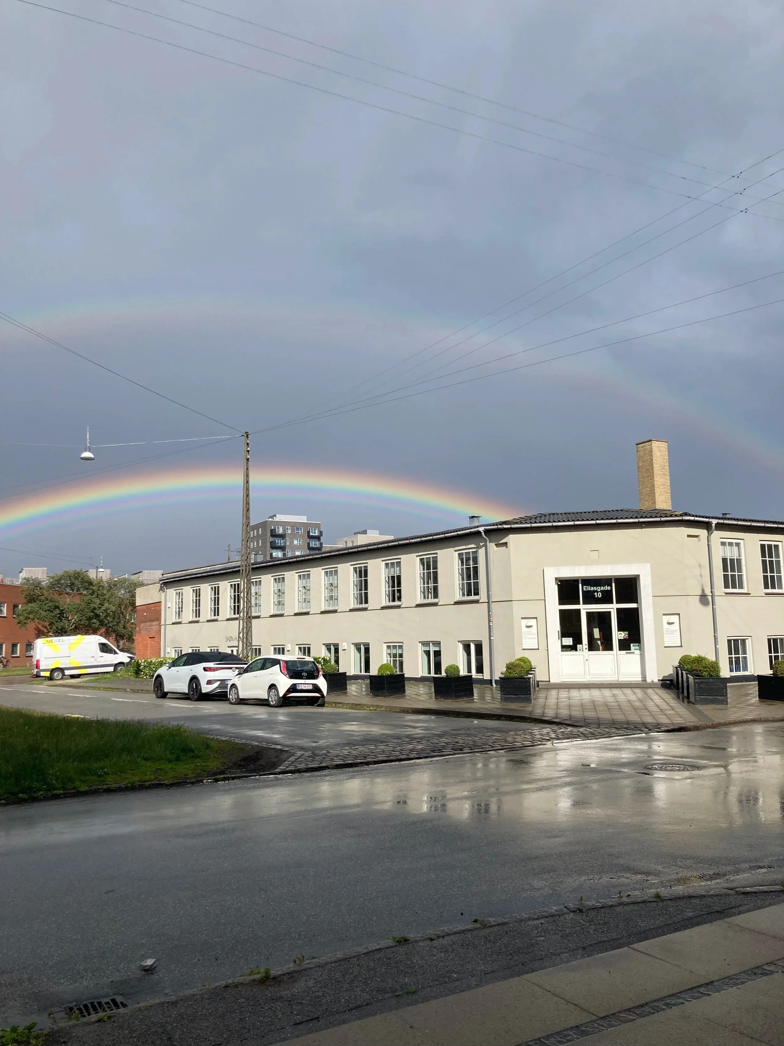 En gade med parkerede biler foran en hvid bygning med vinduer og en indgang, regnvejrspletter på vejen, og en smuk regnbue på himlen.