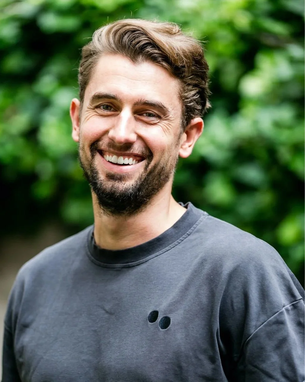 A man with brown hair and a beard smiling outdoors, wearing a gray T-shirt with small black dots on the chest.