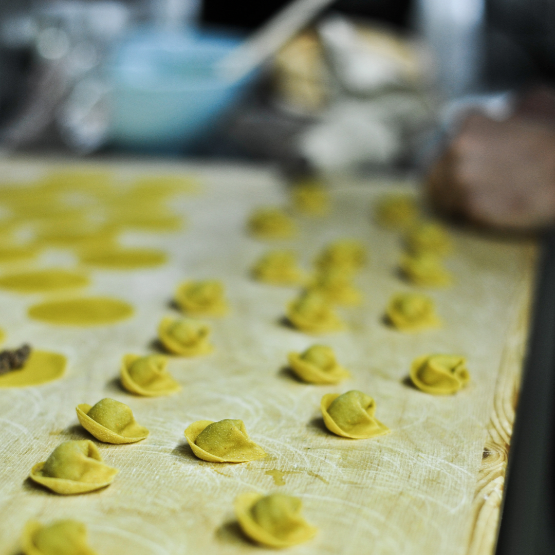 Tortellini verts crus sur une surface farinée, en train d'être préparés, avec des ingrédients et ustensiles de cuisine en arrière-plan.