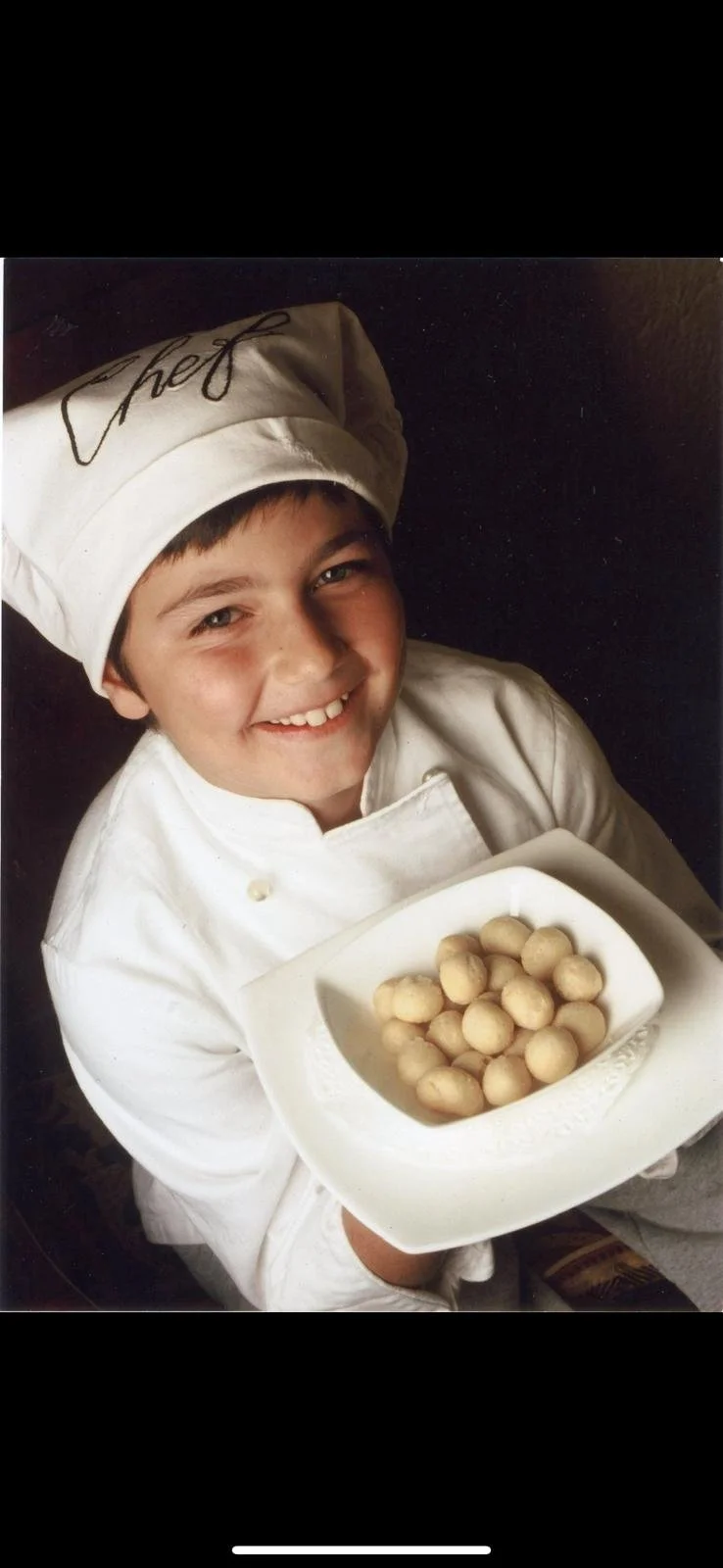 Lorenzo Bucci, petit, en tenu de chef, portant un toque avec le mot 'Chef' écrit dessus, sourit en tenant une assiette blanche avec des petites boules de pâte.