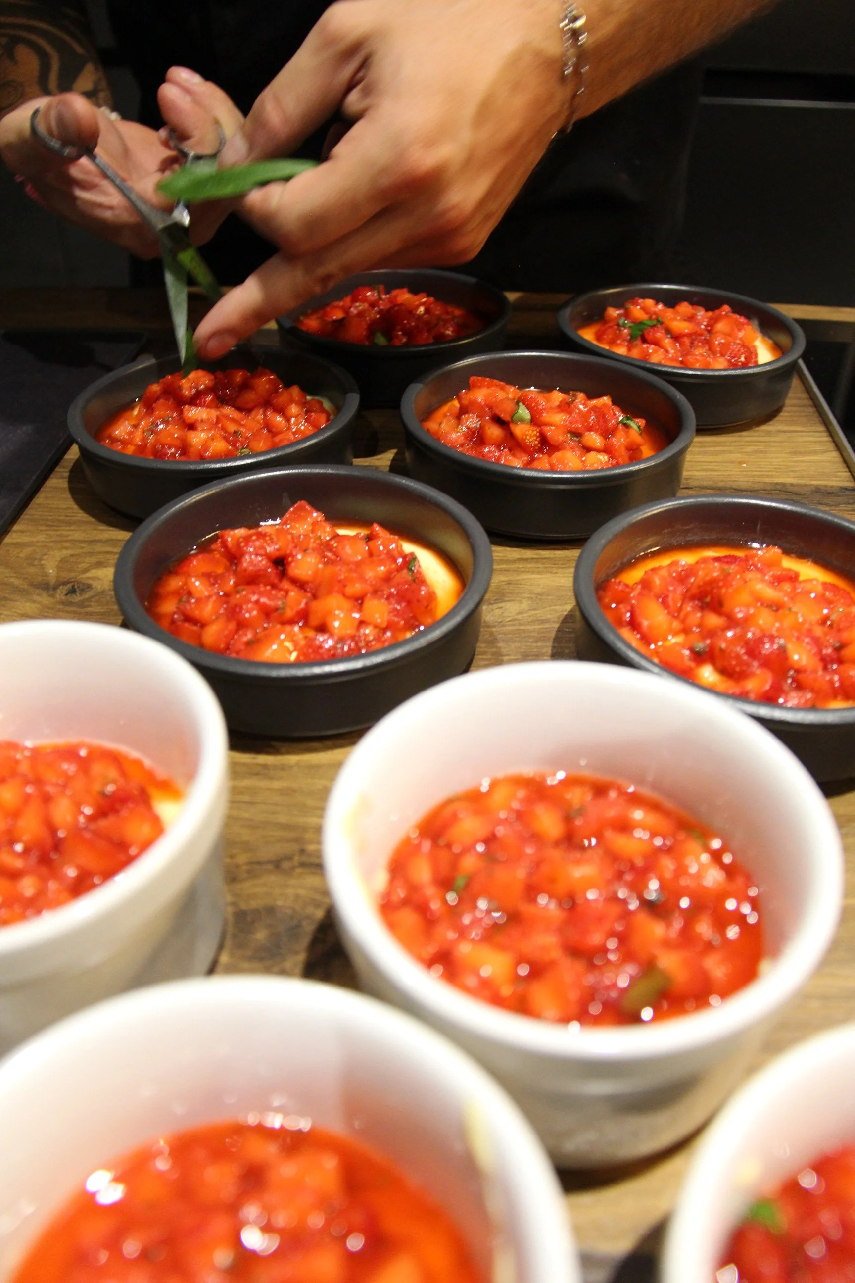 Une personne est en train de garnir des ramequins de fraises & basiliques. La photo est prise en plongée et montre plusieurs bols alignés, avec des mains en train de cuisiner.