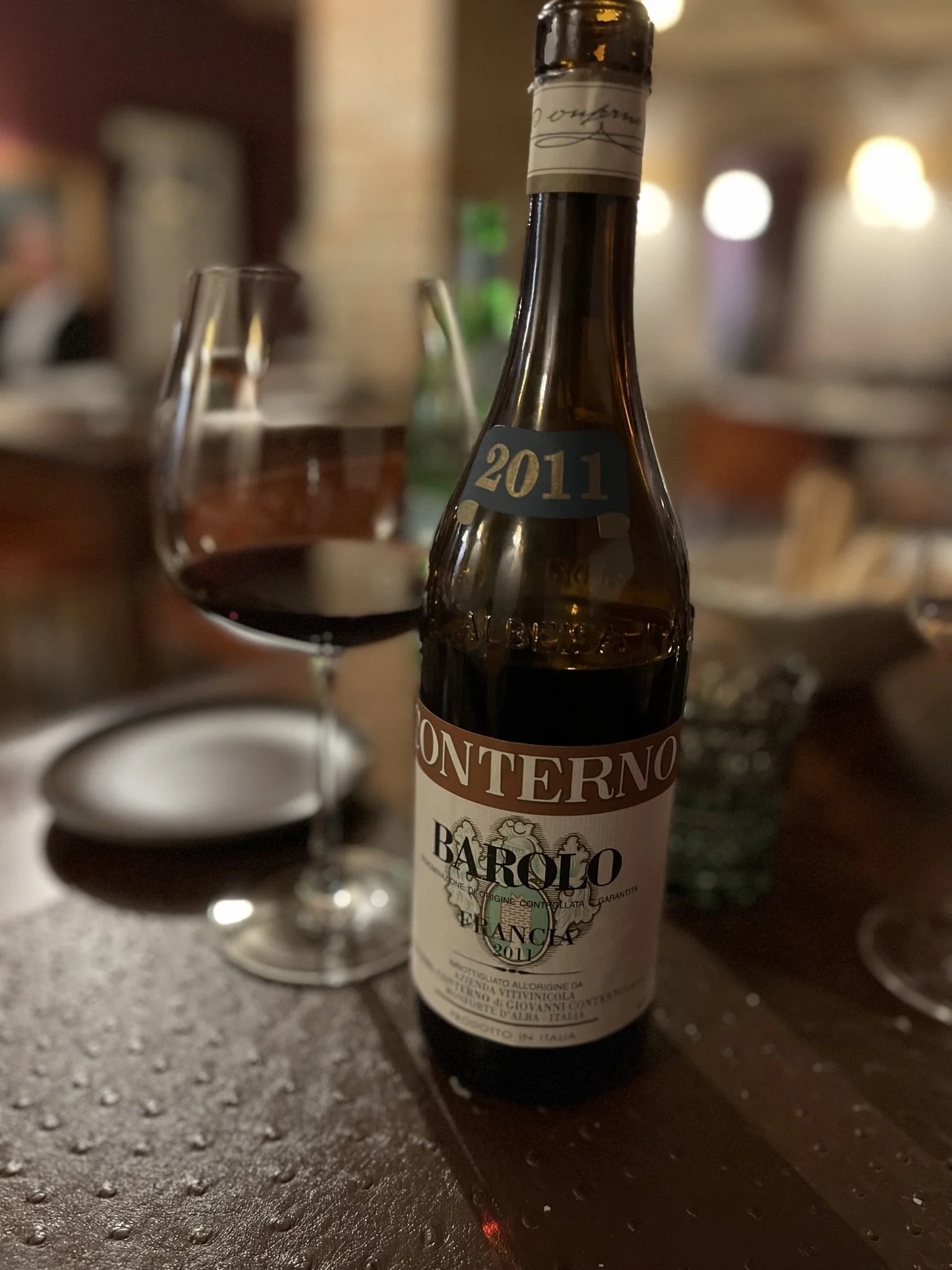 Bouteille de vin rouge de 2011 d'Italie, posé sur une surface en bois, avec un verre de vin rouge flou devant. Le fond est flou avec des lumières chaudes.
