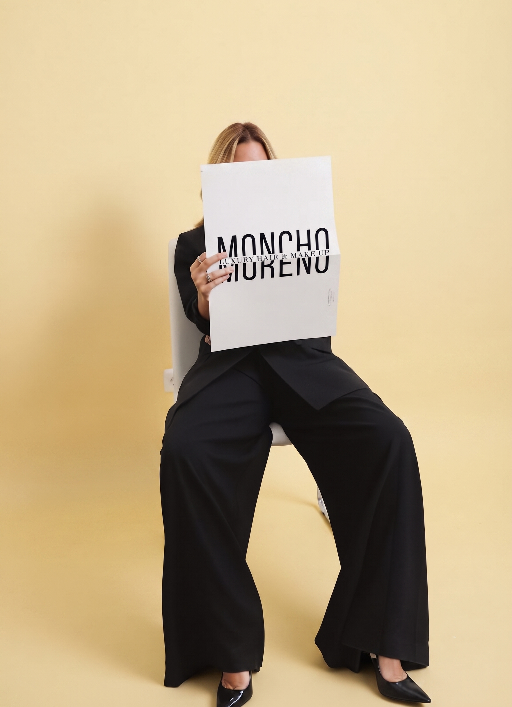 Two women sitting on chairs against a yellow wall, holding a large white sign that reads 'MONCHO GIORGEO LUXURY HAIR & MAKE UP,' with only their head and legs visible.