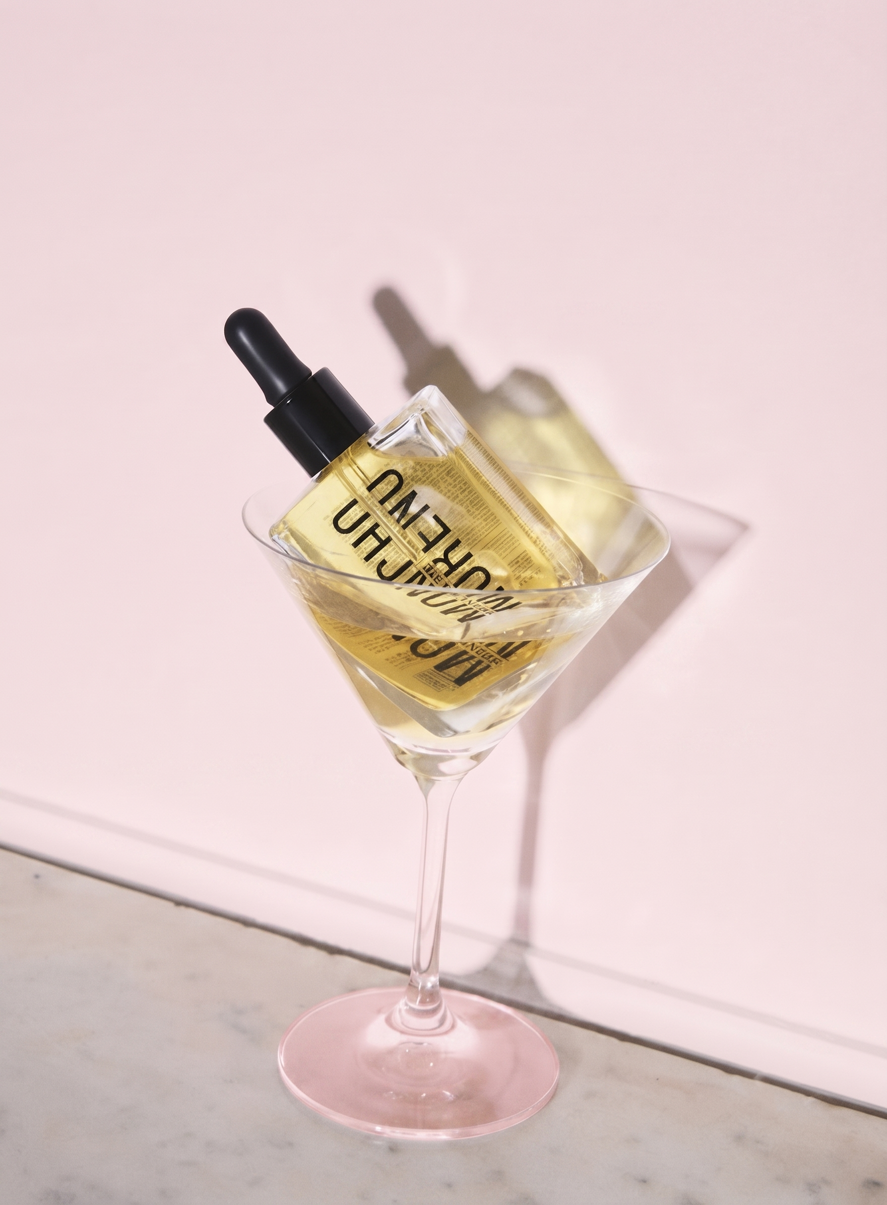 A martini glass with a pink tint, holding a bottle of yellowish oil with a black dropper cap, tilted inside the glass against a light pink background.