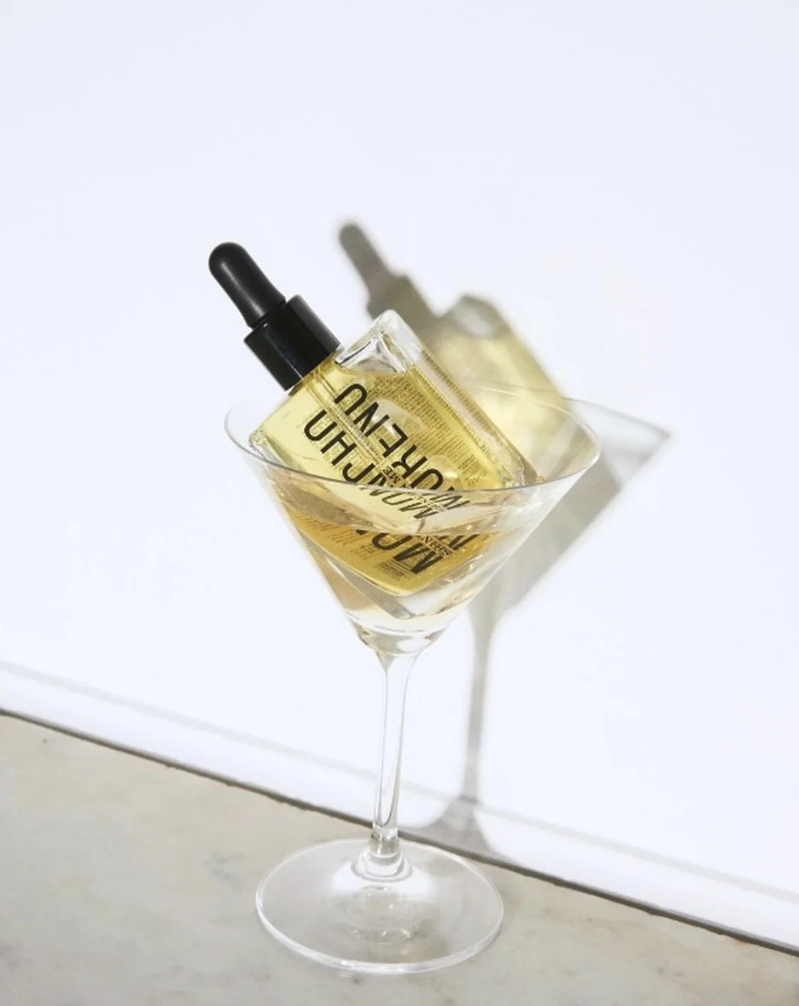 A clear martini glass filled with yellow liquid and a rectangular perfume bottle inside with a black dropper cap. The perfume bottle has text on it, and its shadow is cast on the white background.