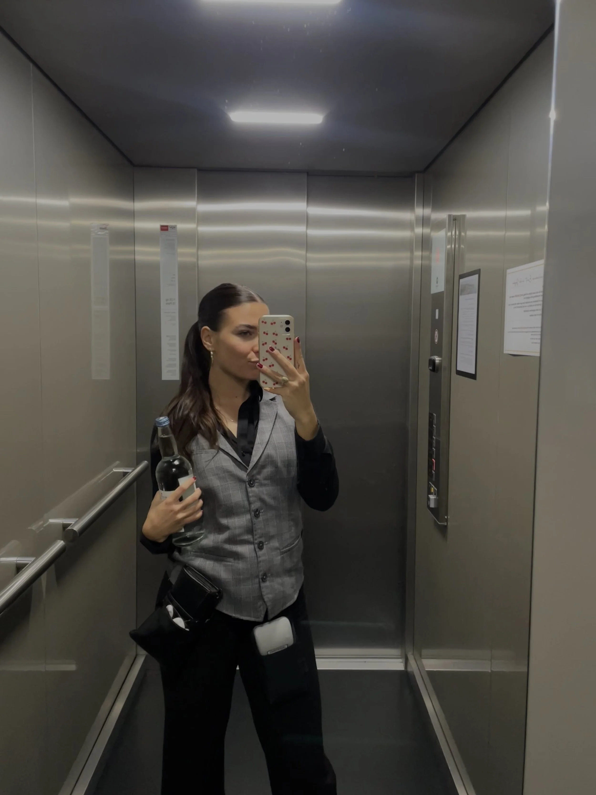 A woman is taking a selfie in an elevator mirror, holding a water bottle and wearing a gray blazer with black pants. She has long, dark hair and is in an elevator with metallic walls and control panel.