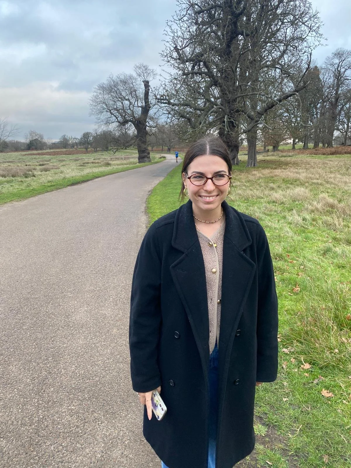 A woman stands on a winding paved path in a park with leafless trees during cloudy weather, smiling and holding a phone.