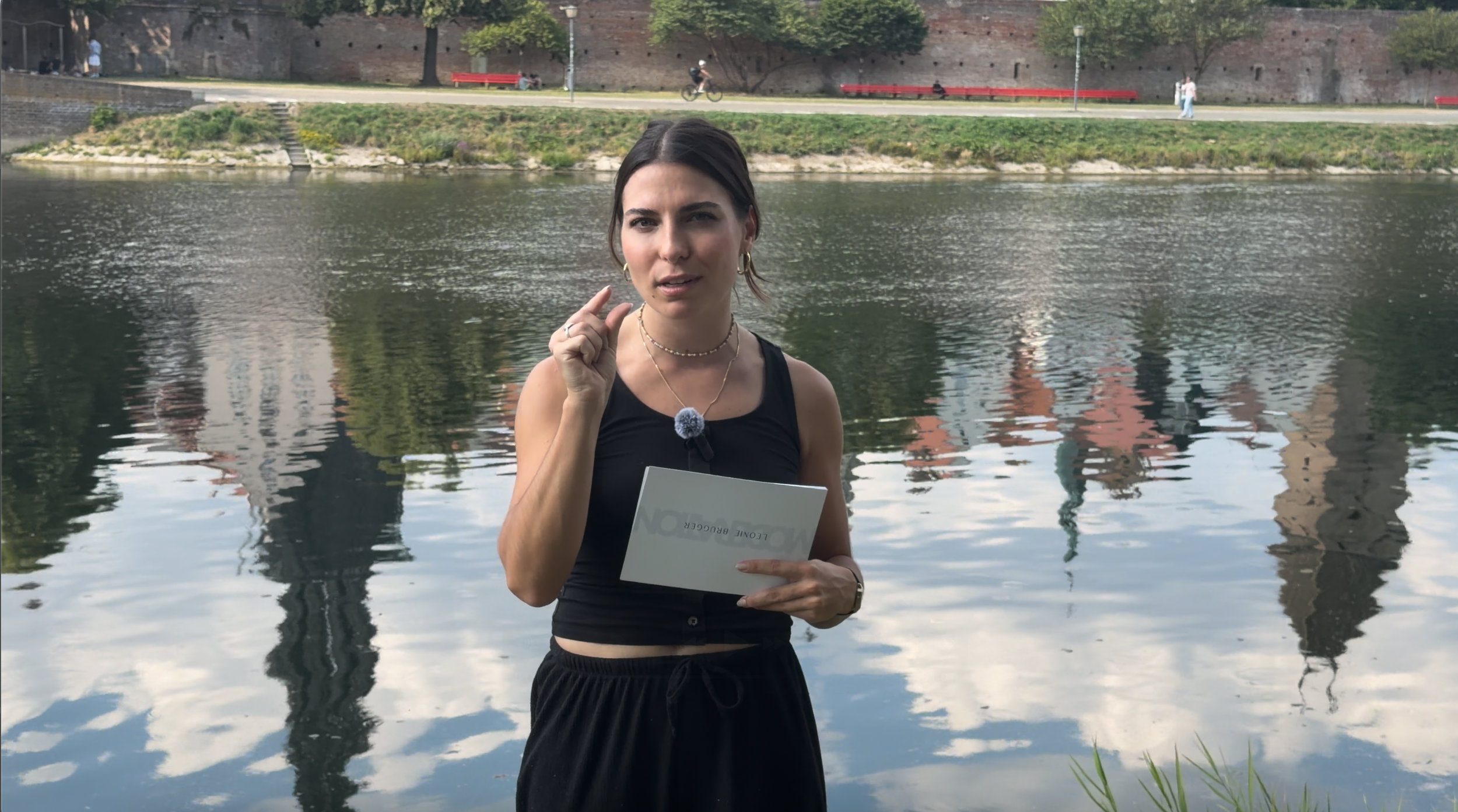 A woman standing near a body of water holding a booklet, with trees, a brick wall, and several people and bicycles in the background.