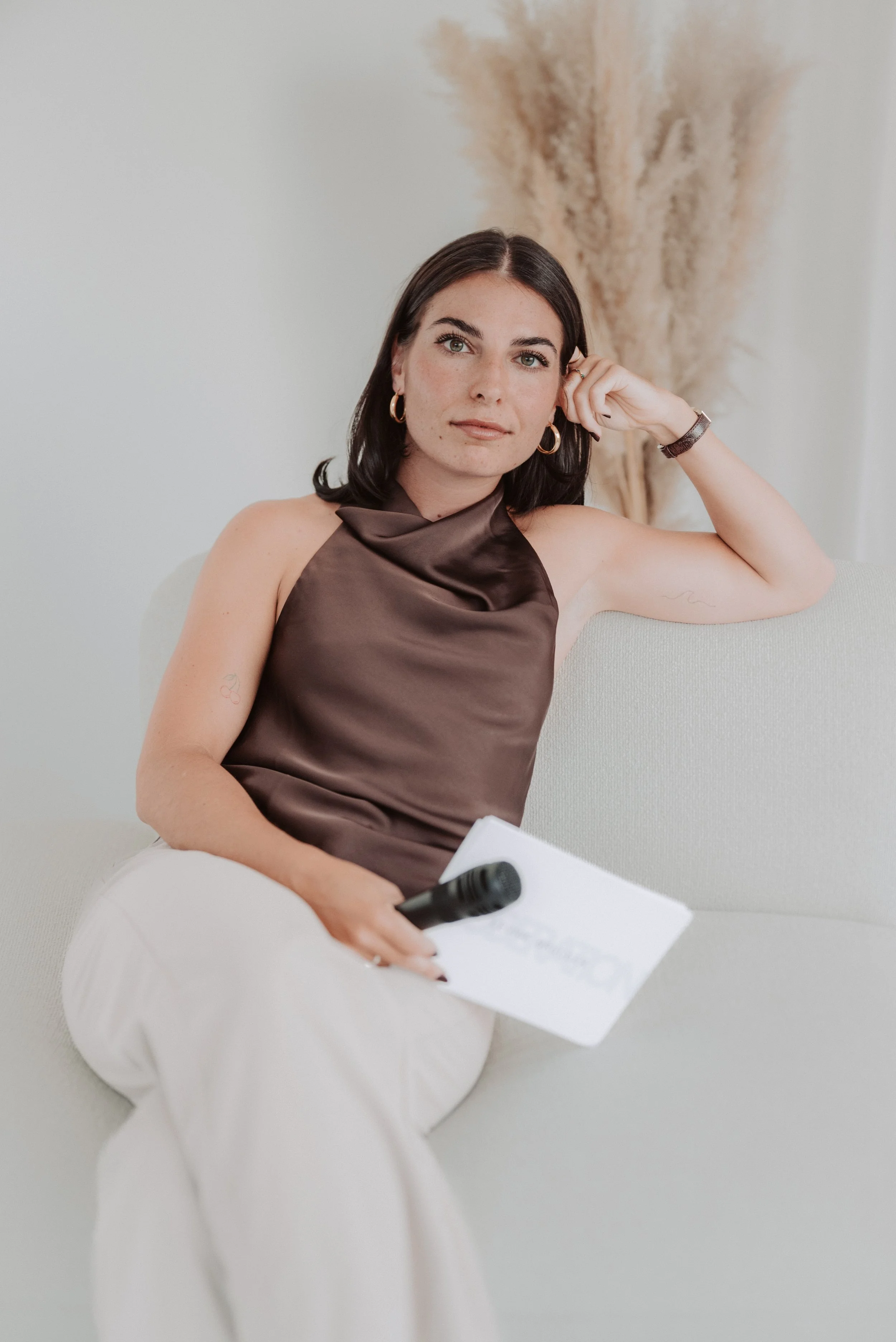 A woman with dark hair and earrings sitting on a light-colored sofa, holding a notepad and microphone, wearing a brown satin top and white pants, with pampas grass decor in the background.