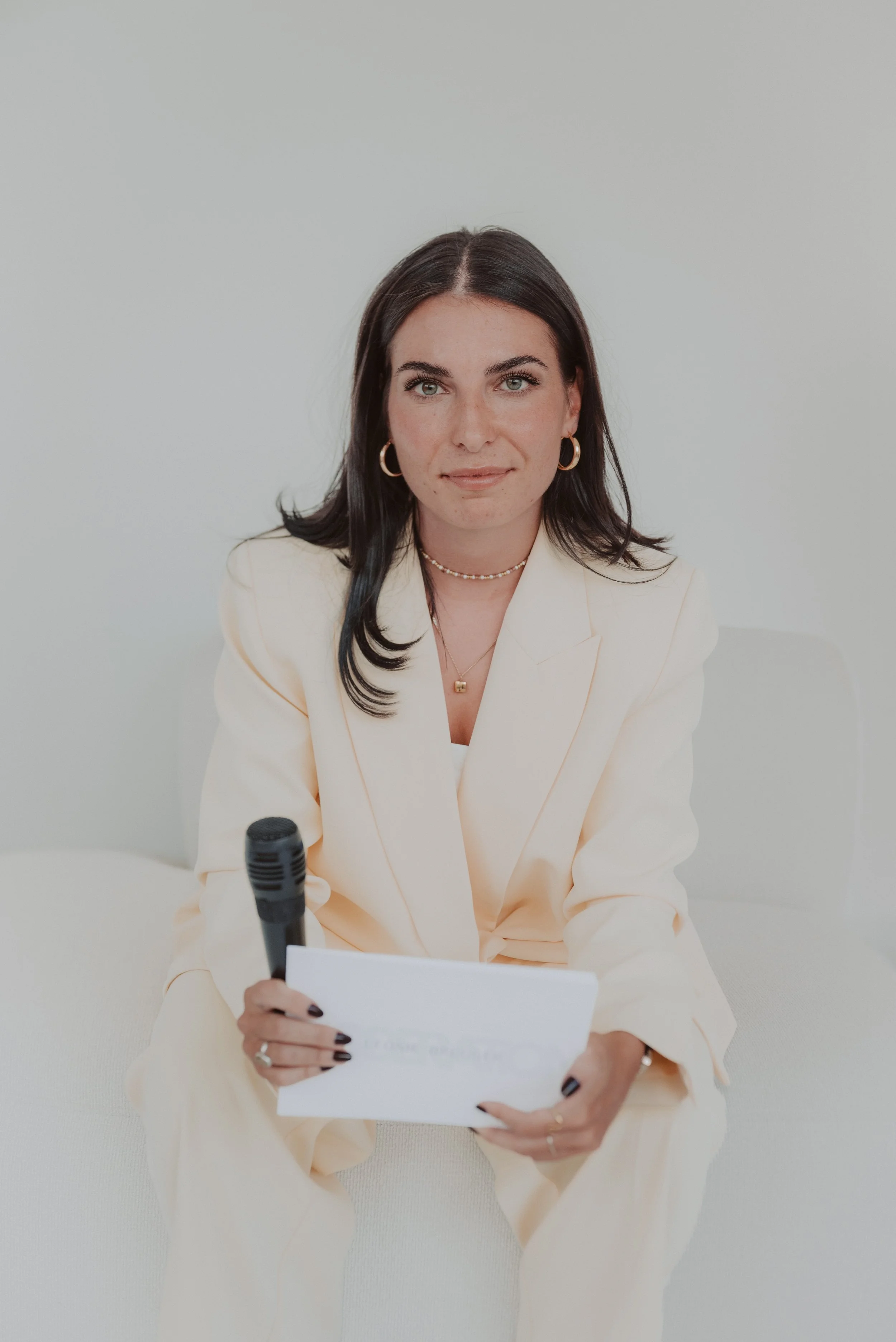A woman with dark hair wearing a cream blazer and gold jewelry, holding a piece of paper and a microphone, sitting against a white wall.