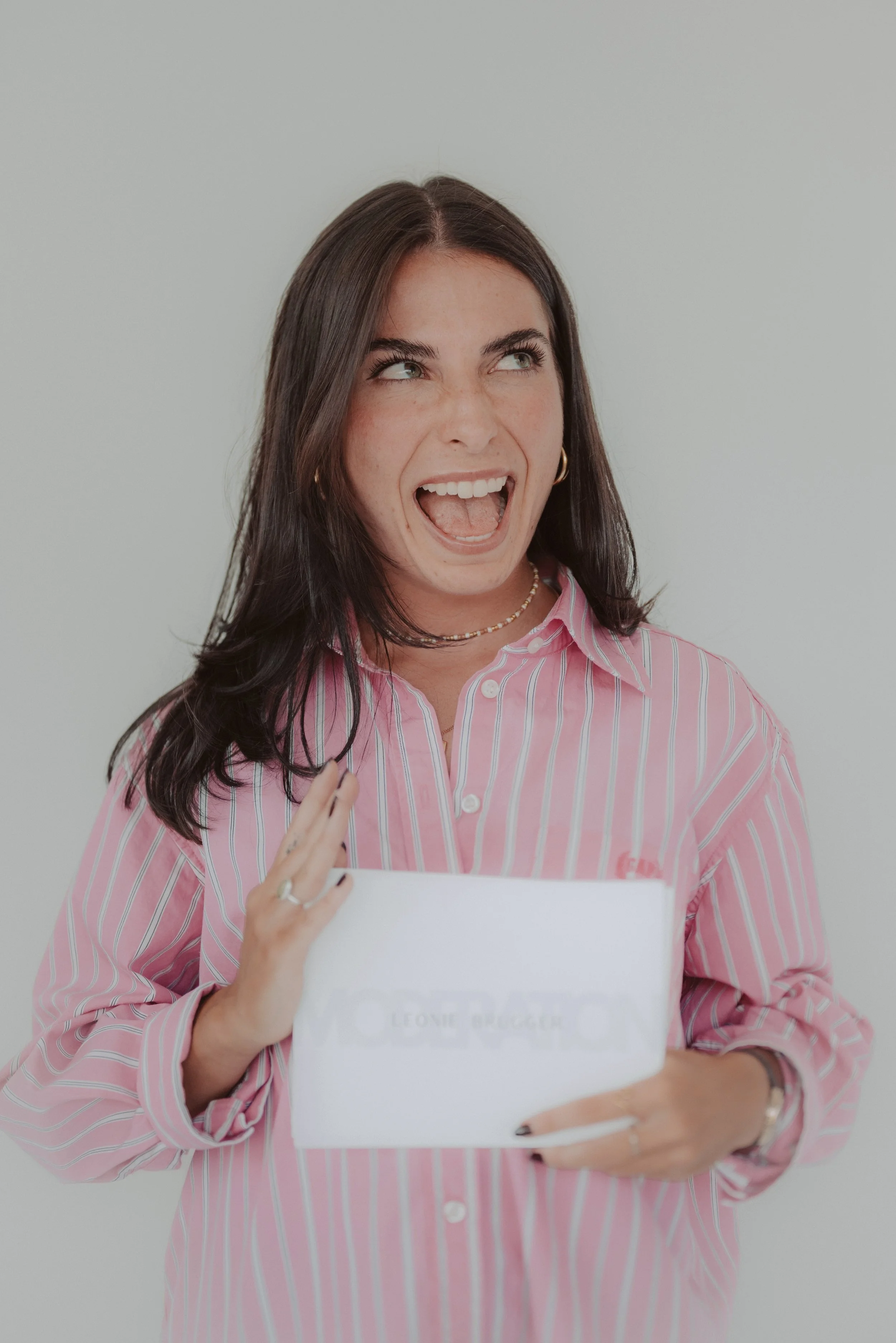 A woman with dark hair, wearing a pink and white striped shirt, holding a white folder, smiling and looking upward.