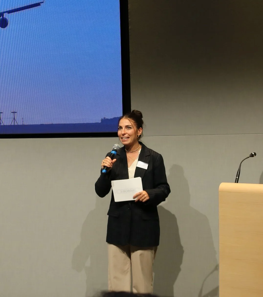 A woman with dark hair in a bun, wearing a black blazer and beige pants, holding a microphone and a piece of paper, speaking at a presentation or conference.