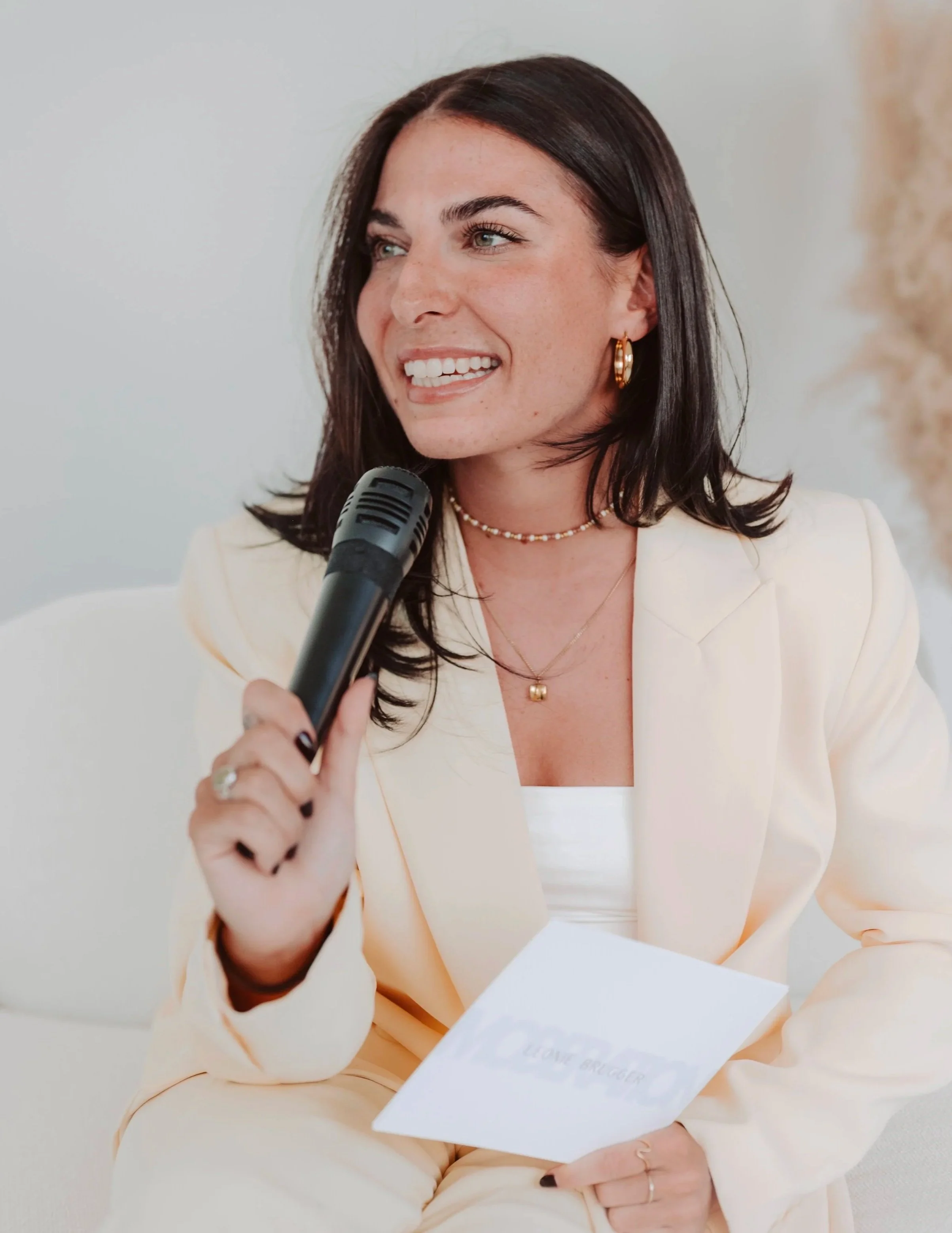 A woman with dark brown hair, wearing gold hoop earrings, a necklace, and a beige blazer, speaking into a microphone while holding a piece of paper.