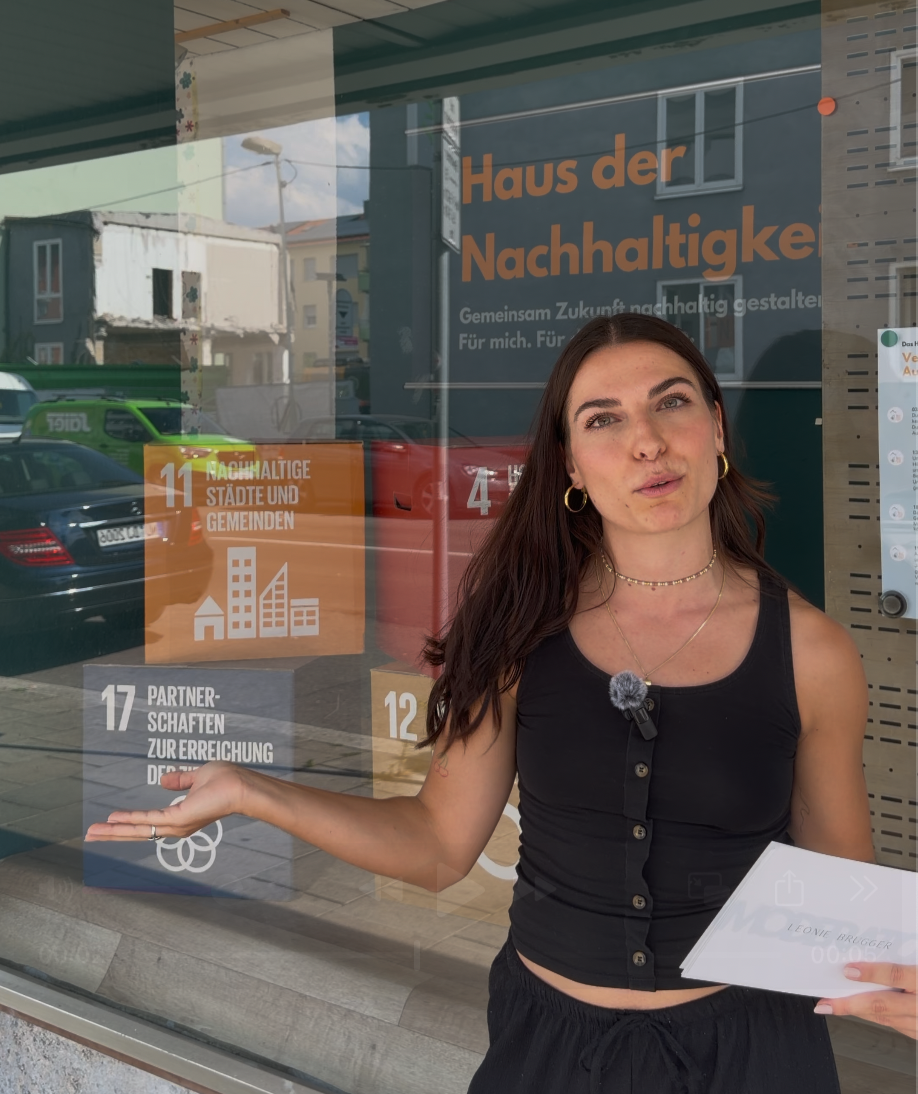 Young woman with long dark hair, wearing a sleeveless black top and jewelry, standing outside a glass window with a reflection of street and parked cars, gesturing with her left hand, holding paper with her right hand, in front of a sign about sustainability.