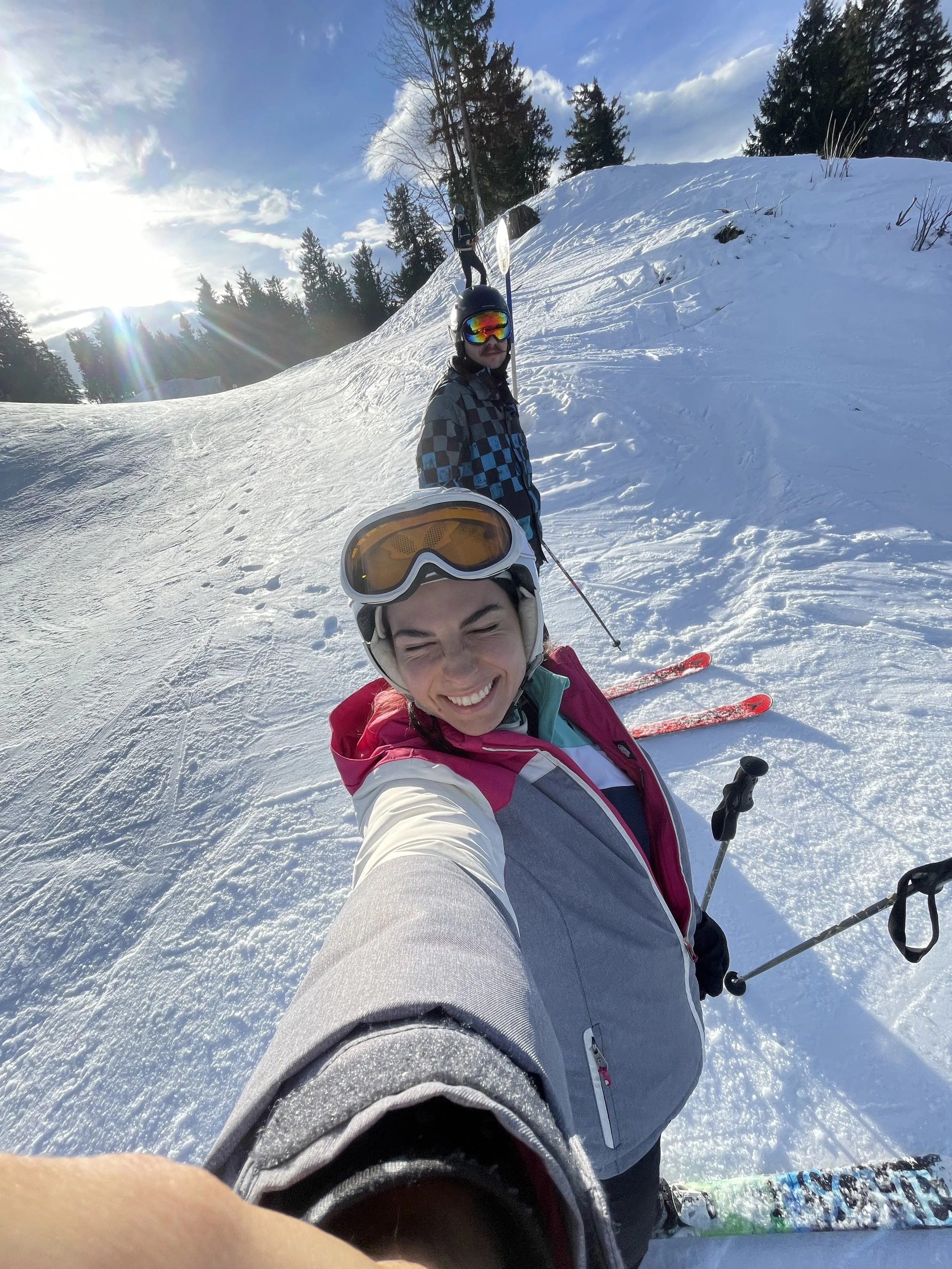 Three people in ski gear, smiling, on a snow-covered mountain with trees and a partly cloudy sky in the background.