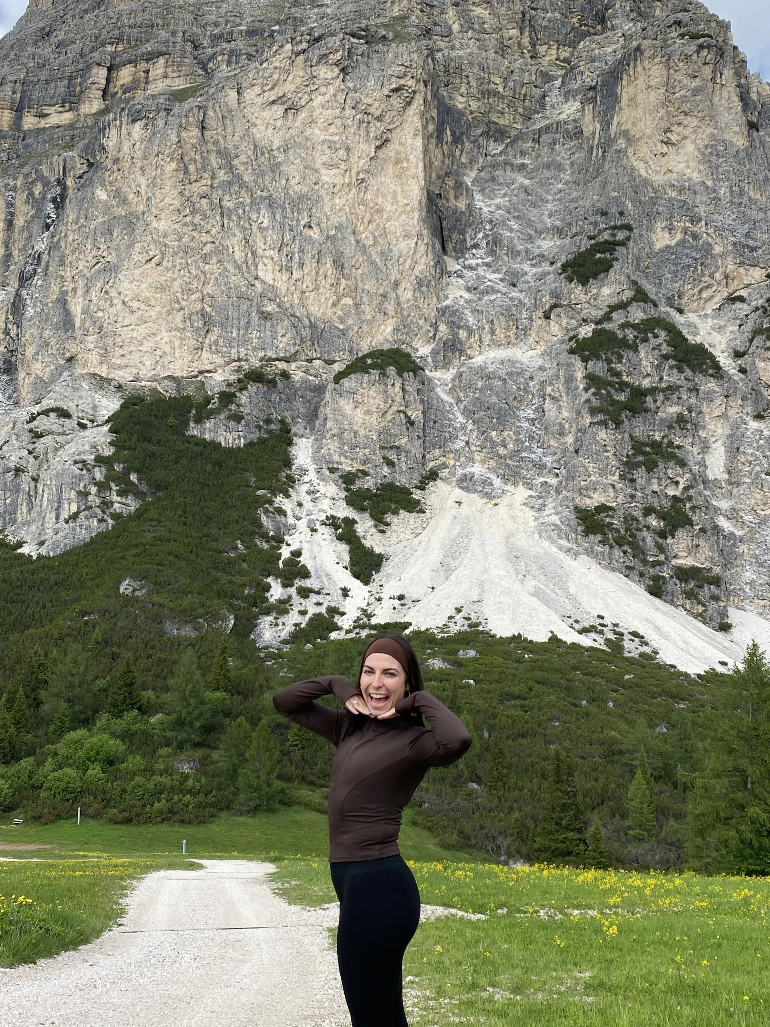 A woman smiling and posing with her hands near her face in front of a mountain, greenery, and a gravel path.