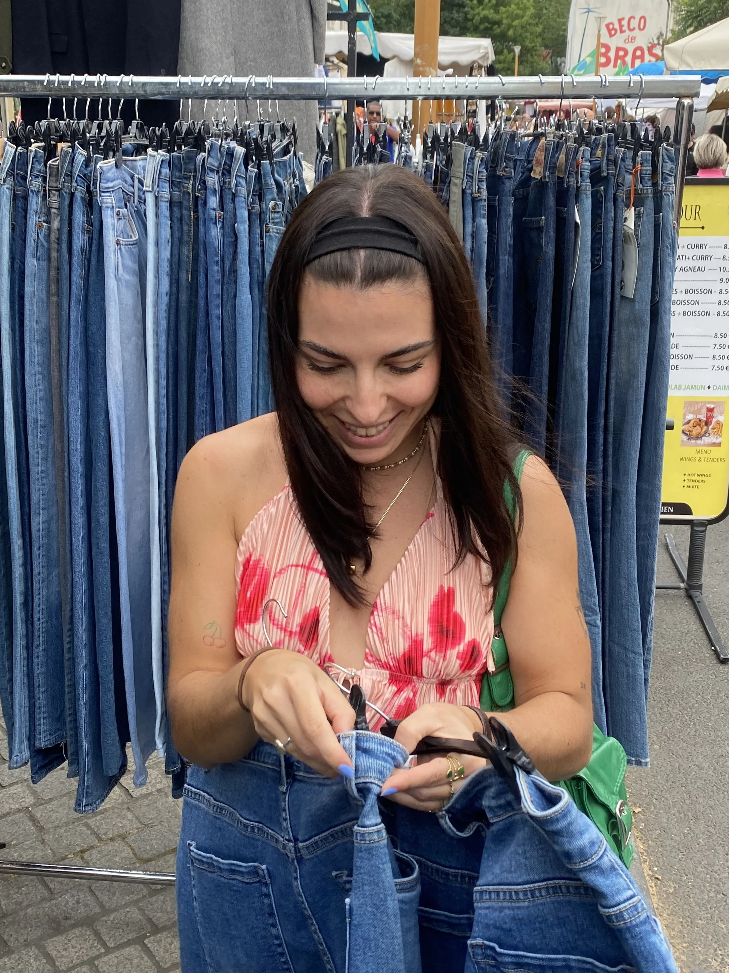 A woman with dark hair, wearing a black headband, a pink and red floral top, and blue jeans, is at an outdoor market. She is smiling and looking down as she adjusts her jeans. Behind her, there is a display of hanging denim jeans and a yellow menu sign with food and drink options. Other market stalls and people are visible in the background.