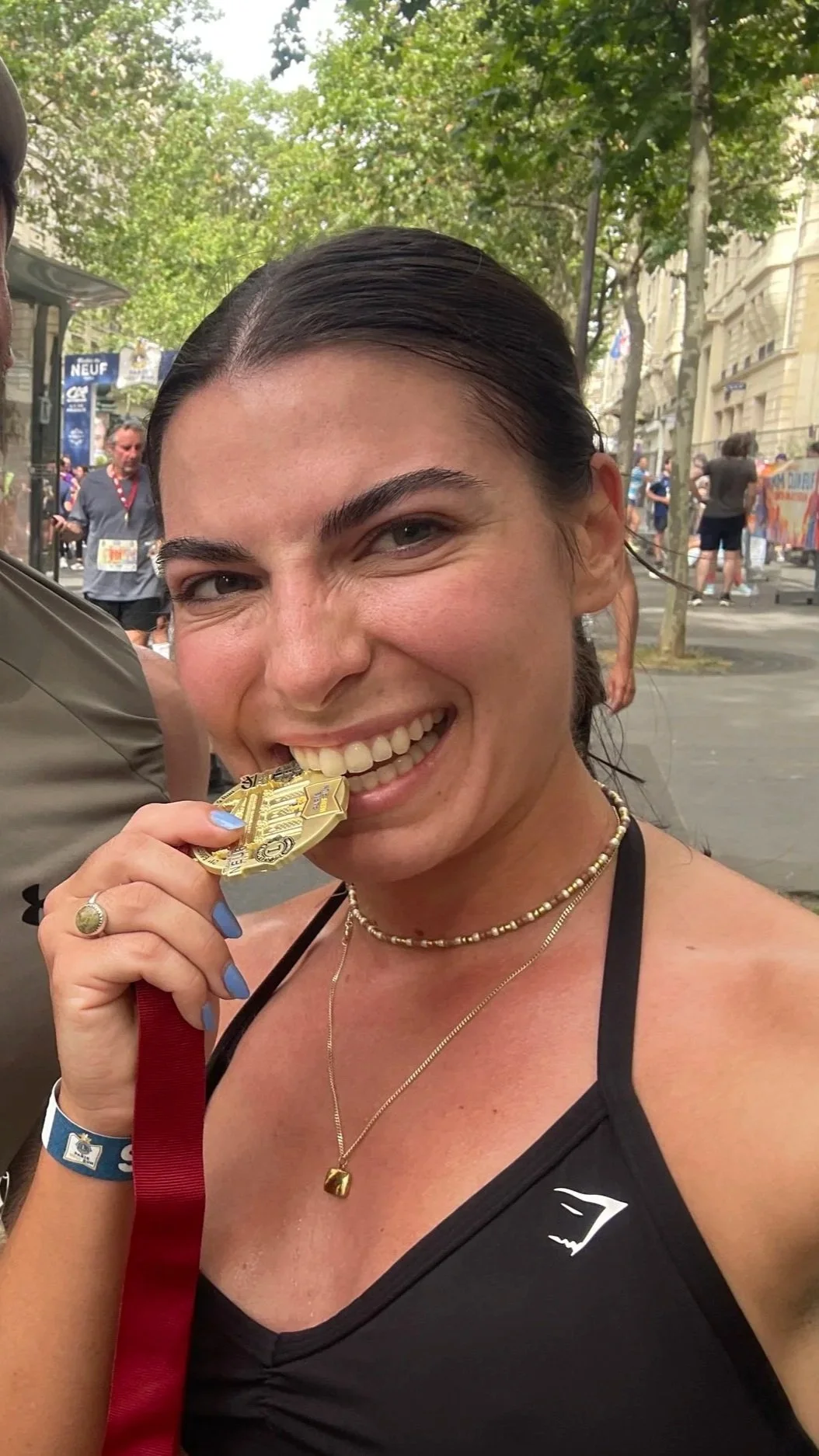 A woman is smiling and biting a finisher medal at an outdoor event, with trees and people in the background.