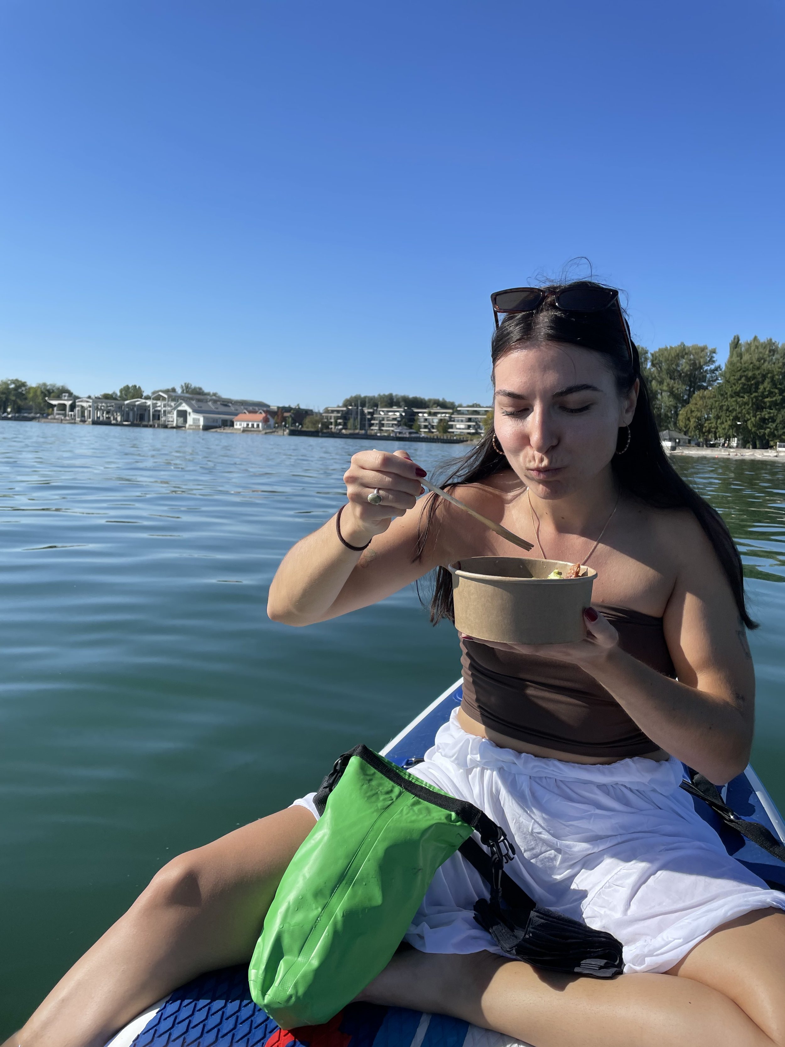 Young woman sitting on a paddleboard, holding a bowl of food, on a lake with buildings and trees in the background.