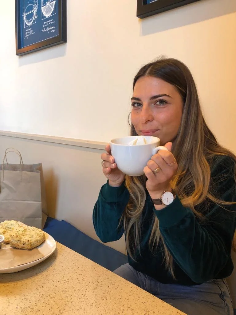 A woman with long brown hair, wearing a dark jacket and a watch, is sitting at a table in a cafe, holding a white mug close to her lips. There is a plate with a scone or biscuit on the table, and a paper bag in the background. She is looking at the camera with a slight smile.