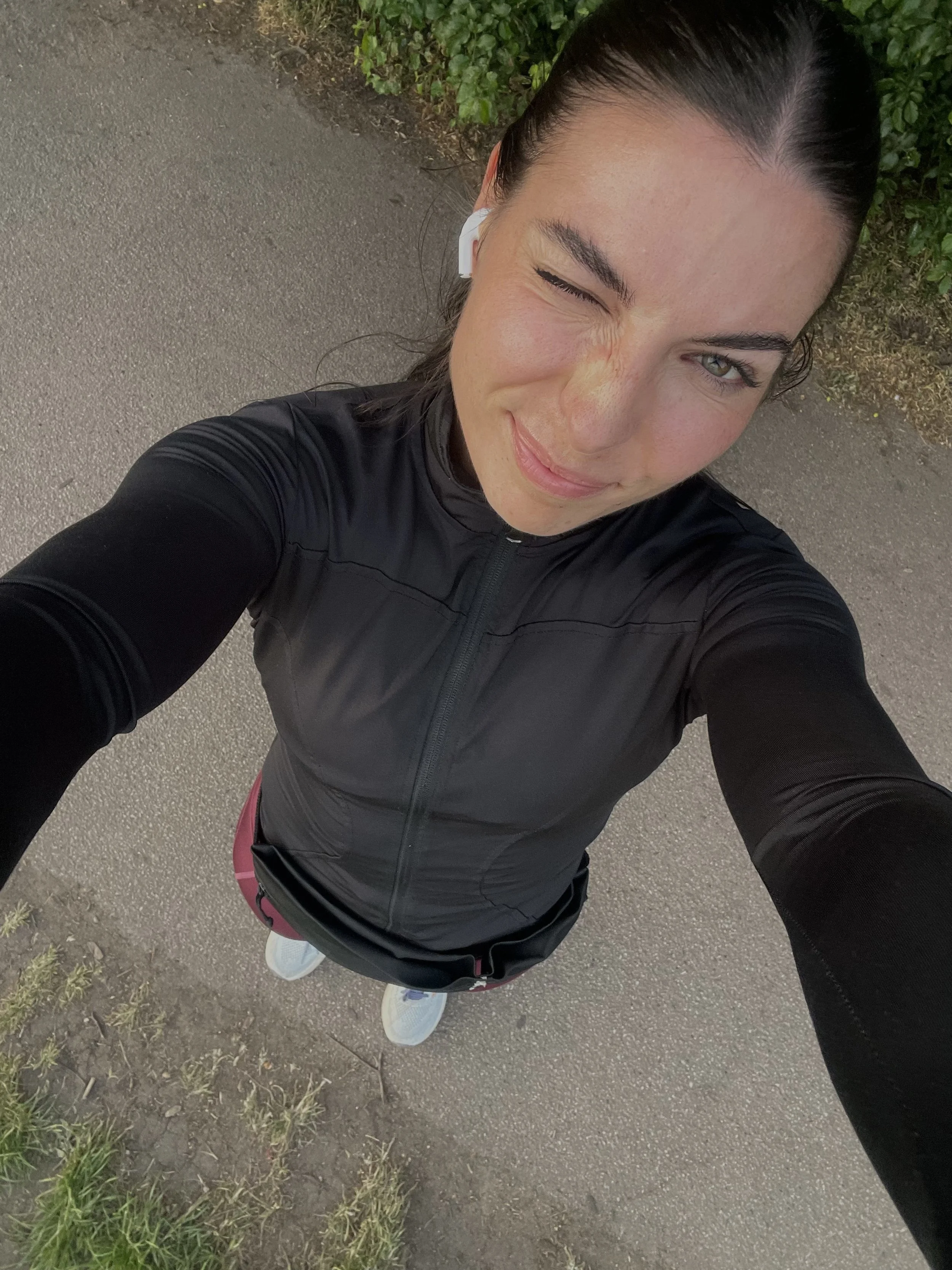 A woman taking a selfie from above while walking outdoors on a paved path, wearing a black athletic jacket and pink shorts, with white sneakers. She has dark hair tied back and is winking at the camera.