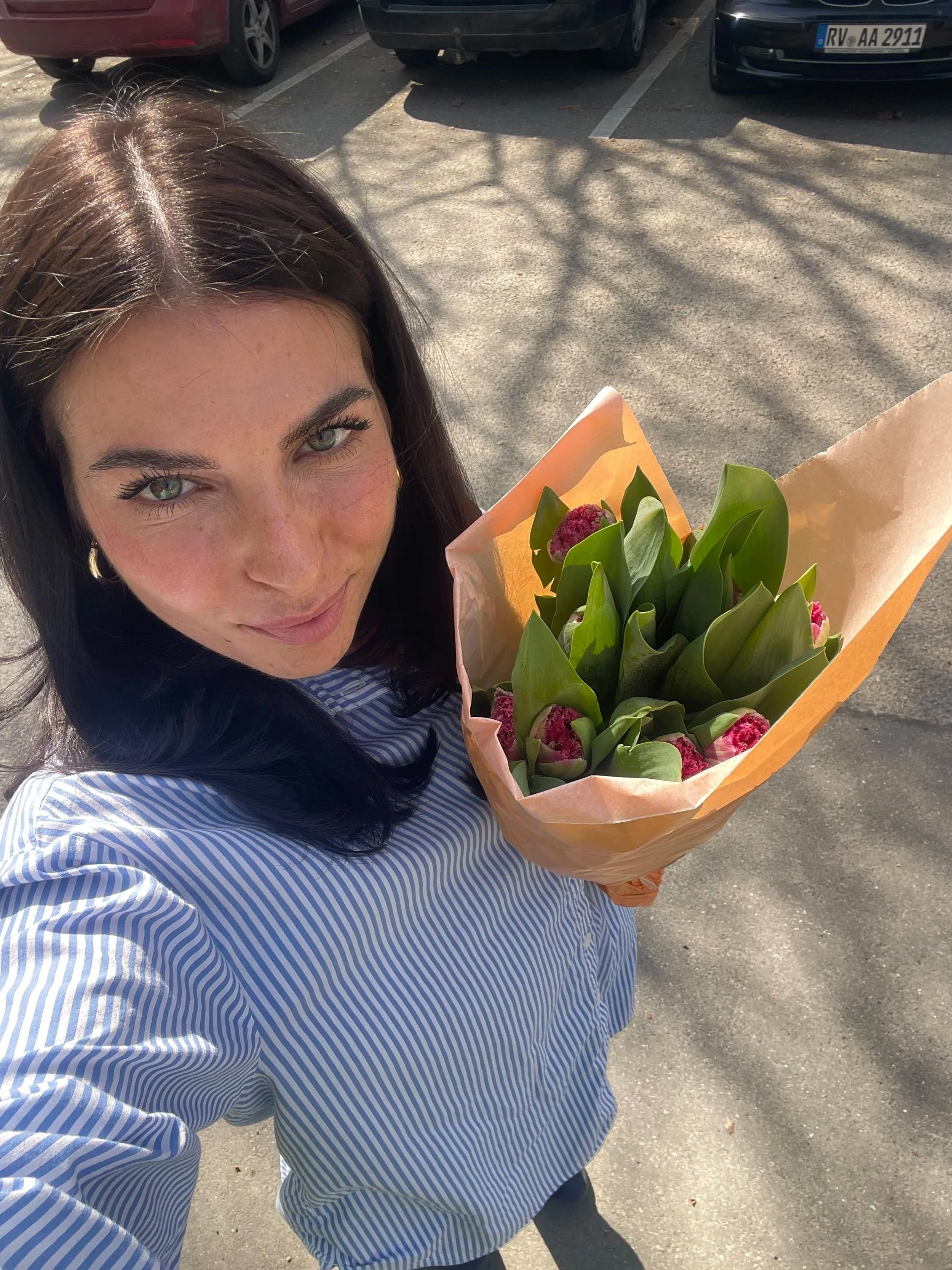 A woman with dark hair and green eyes holding a bouquet of pink tulips in a parking lot on a sunny day.