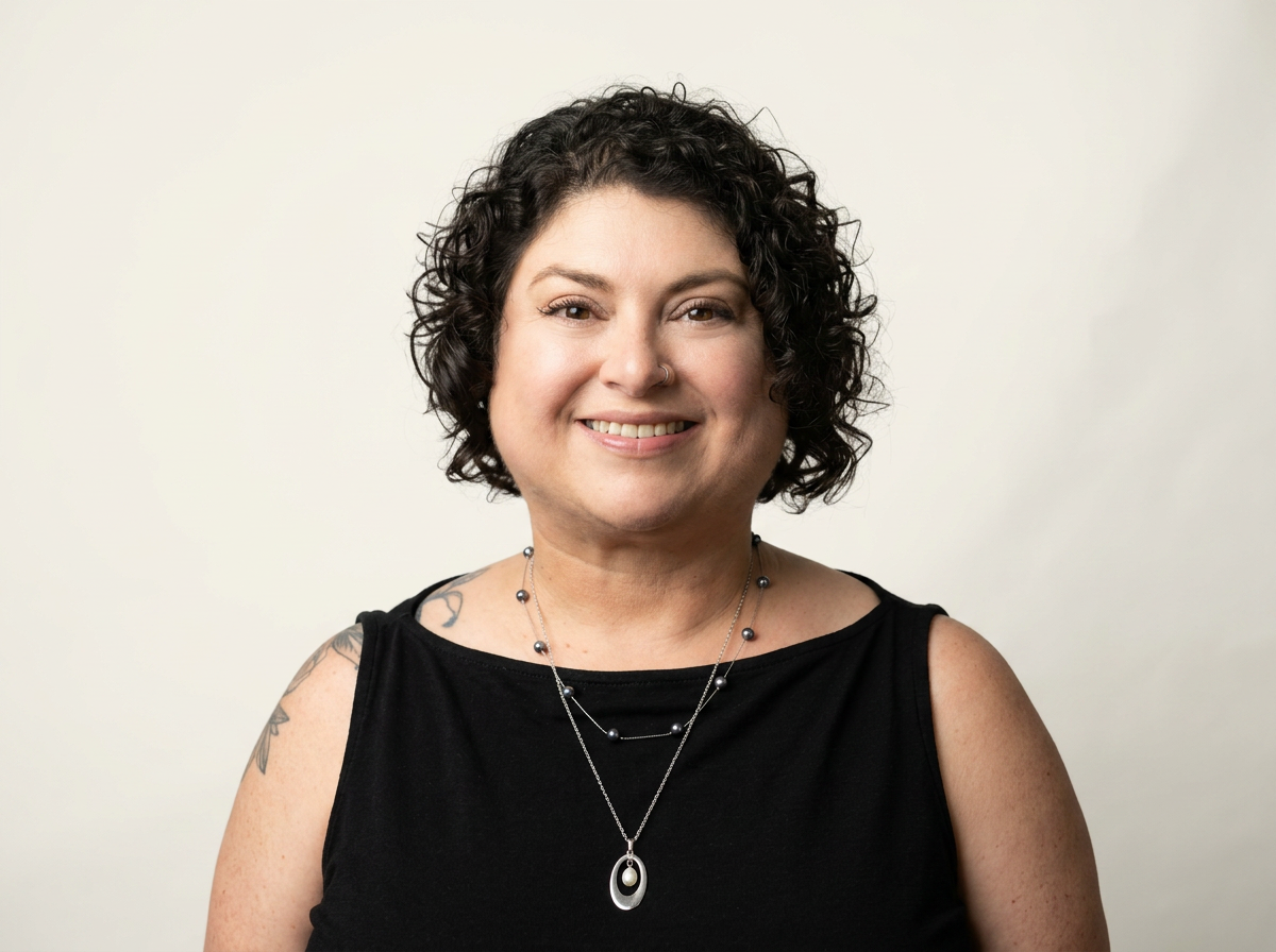 A headshot of Jules, Disorganized Mind founder, wearing a black sleeveless top and two necklaces. Part of a tattoo is showing on her shoulder. She is smiling and has brown eyes and short curly hair.