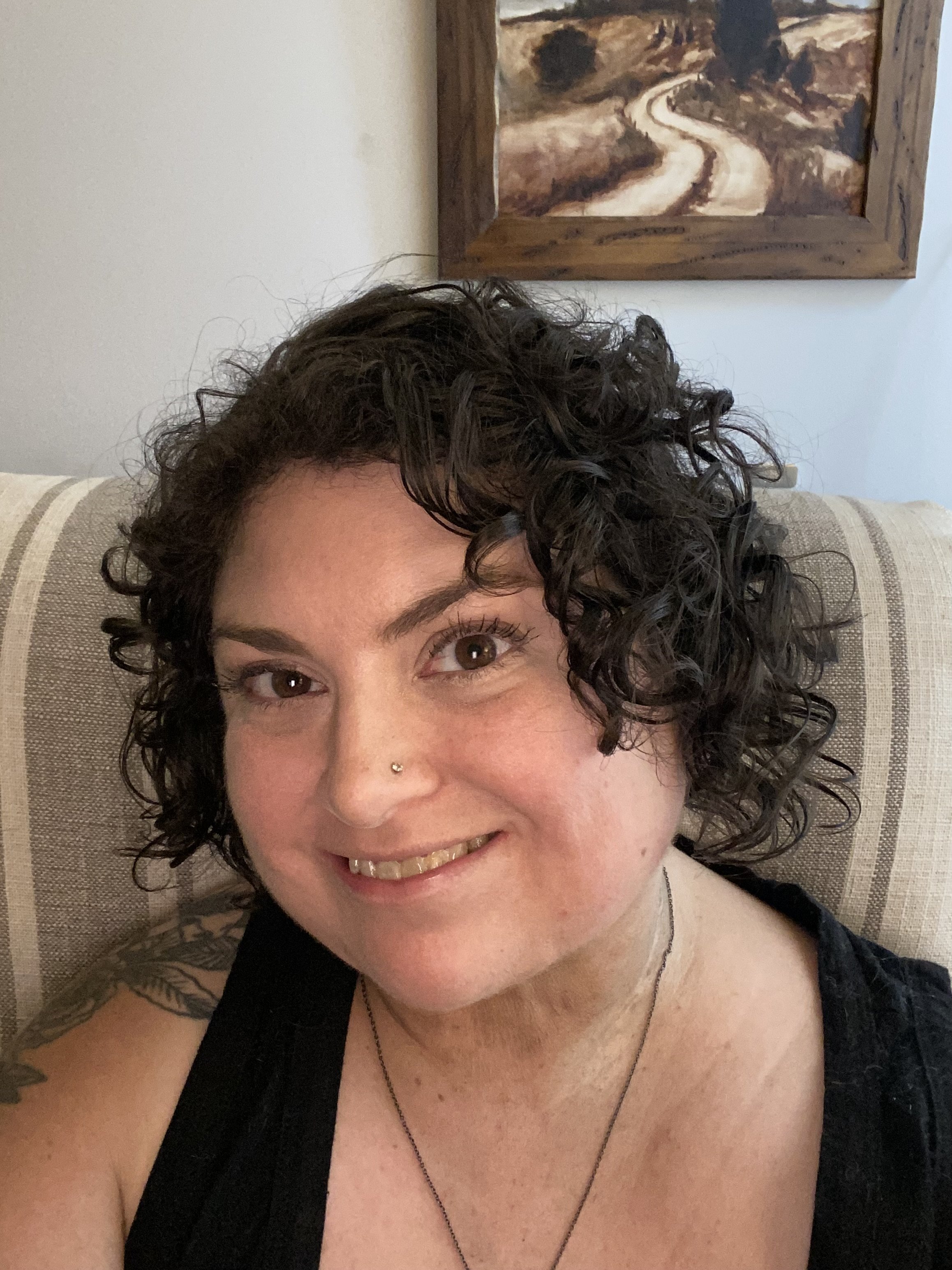 Close-up of Jules with dark curly hair, brown eyes, a nose piercing, smiling, sitting on a beige striped armchair, with a landscape painting on the wall behind her.
