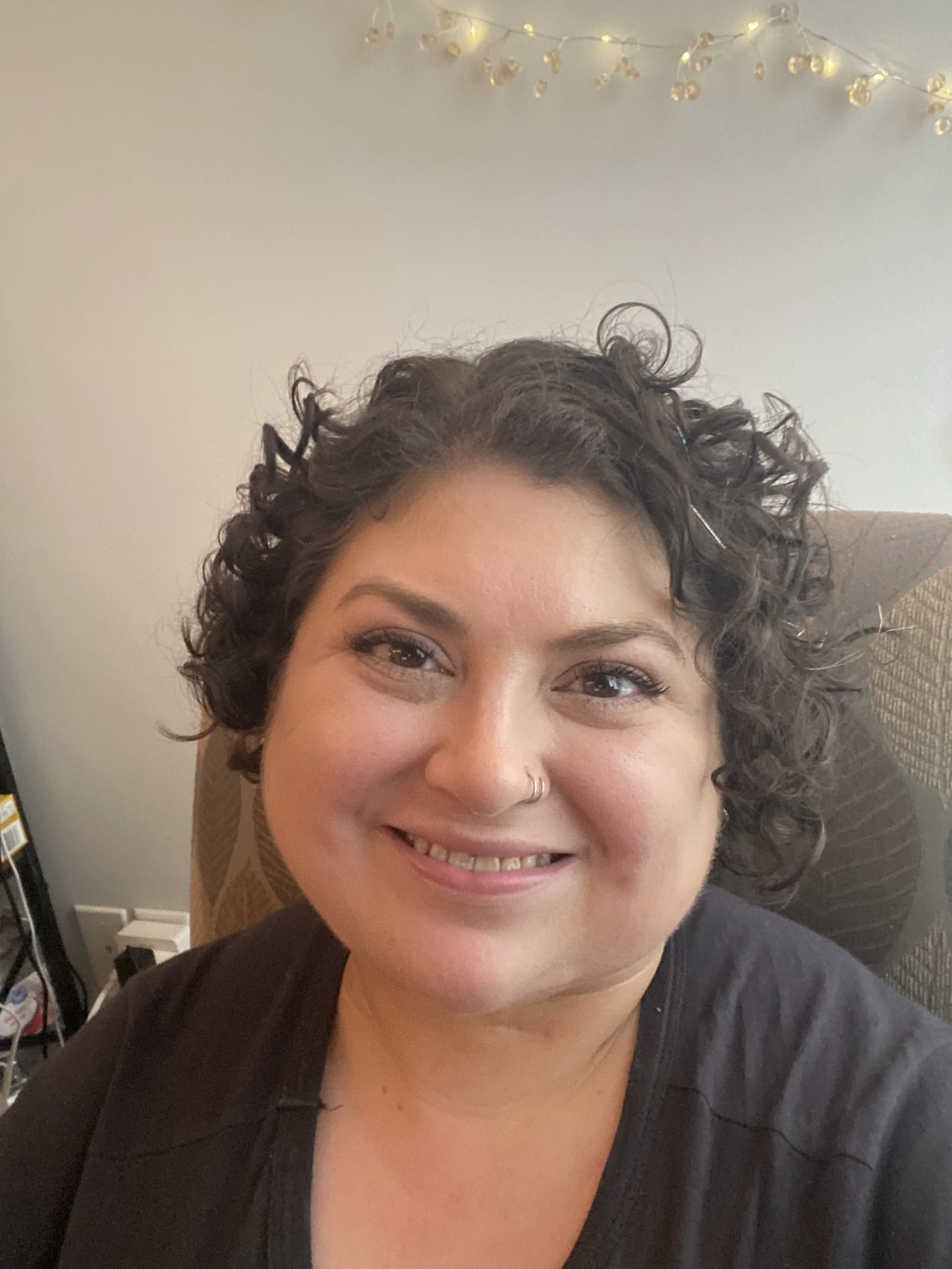 an headshot of Jules wearing a dark gray shirt, sitting in a brown chair with string lights hanging above. She is smiling with her head slightly tilted, and has short, dark brown, curly hair
