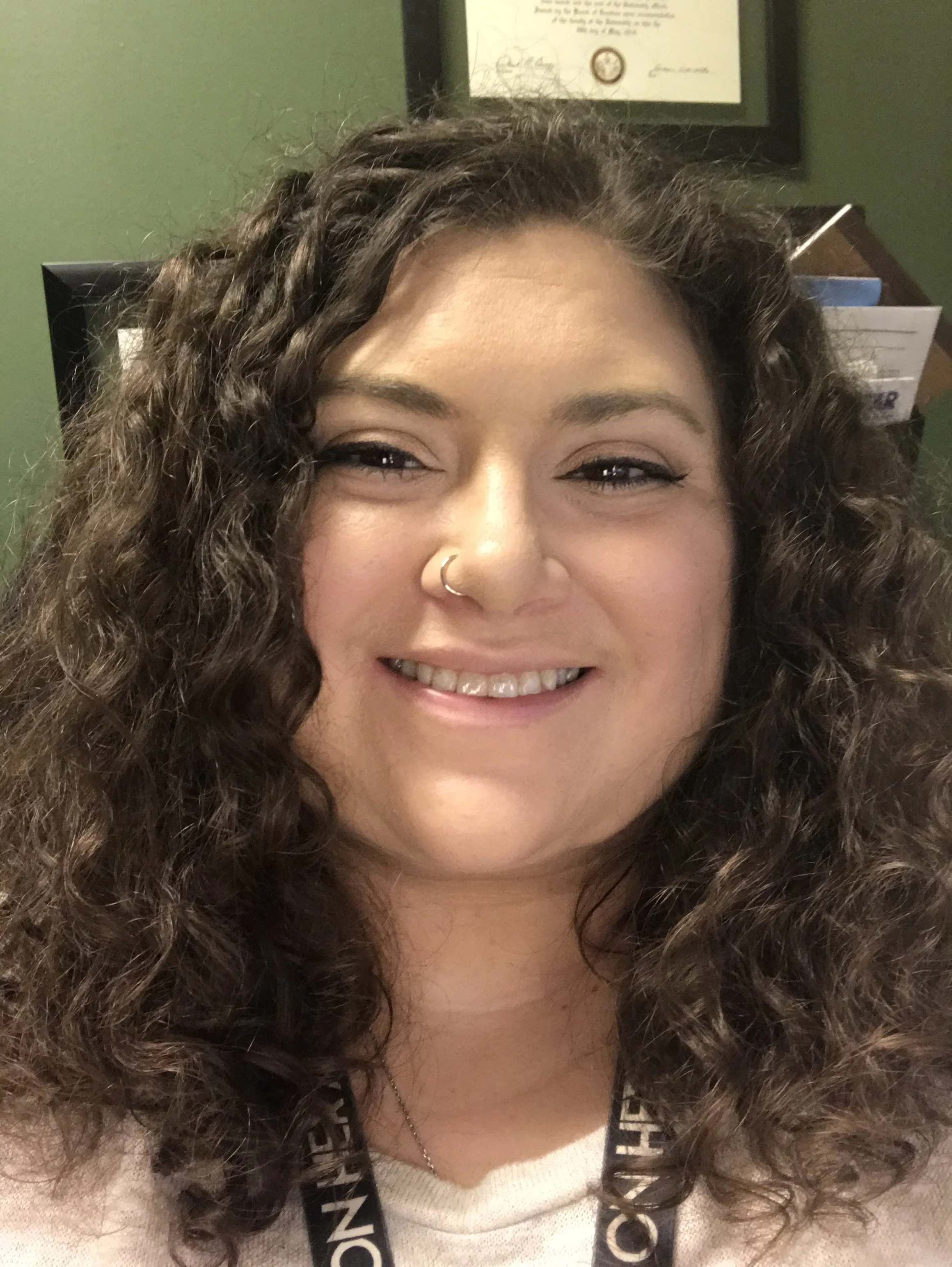 A headshot of Jules, Disorganized Mind creator in a green office with some diplomas behind her head. She is wearing a white shirt with a black lanyard with white text showing. Jules is smiling and has long, dark brown, curly hair