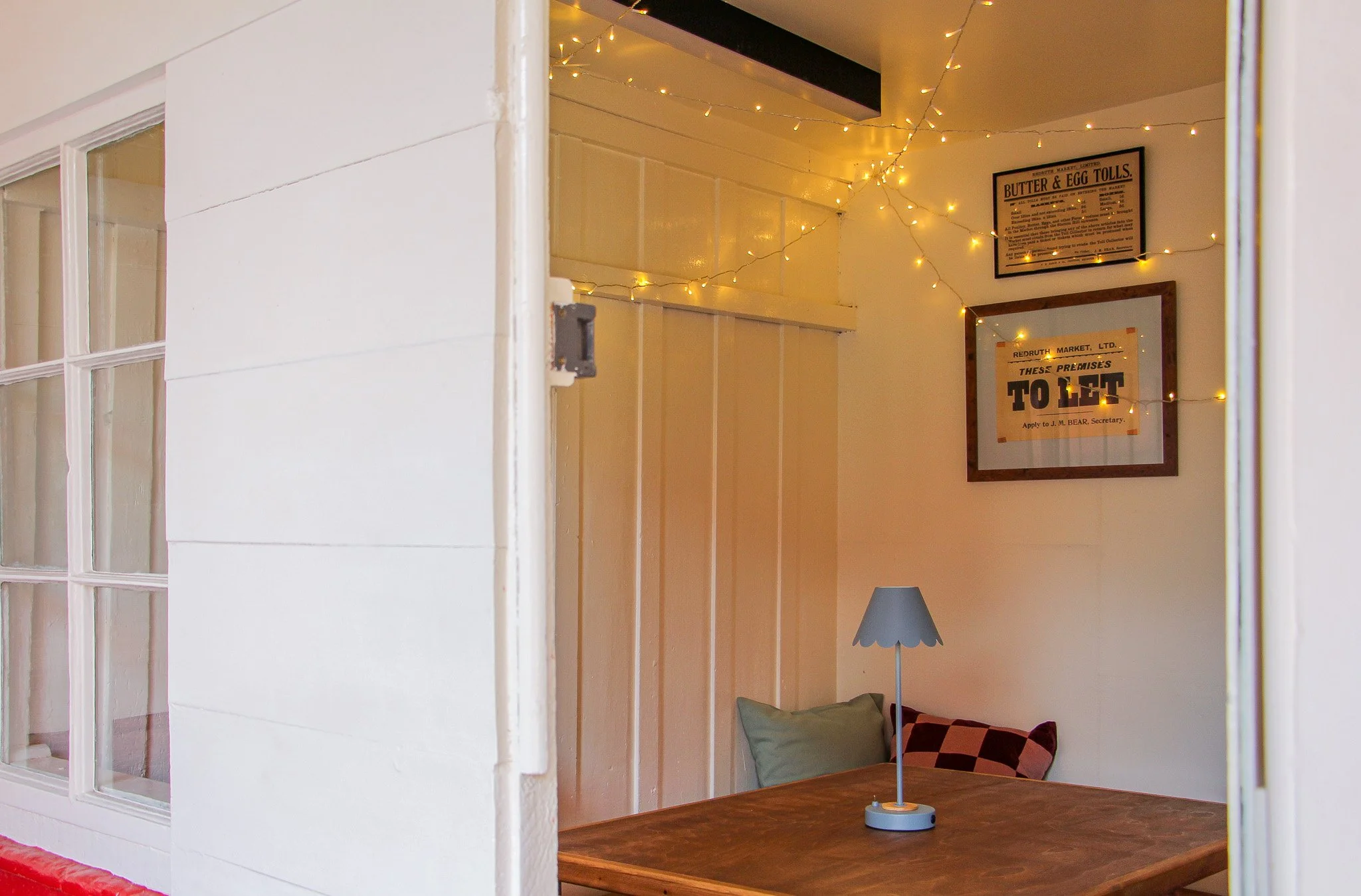 Interior of a cozy room with white walls decorated with string lights. There are framed posters on the wall, a table with a modern lamp, and some cushions on a bench or sofa.
