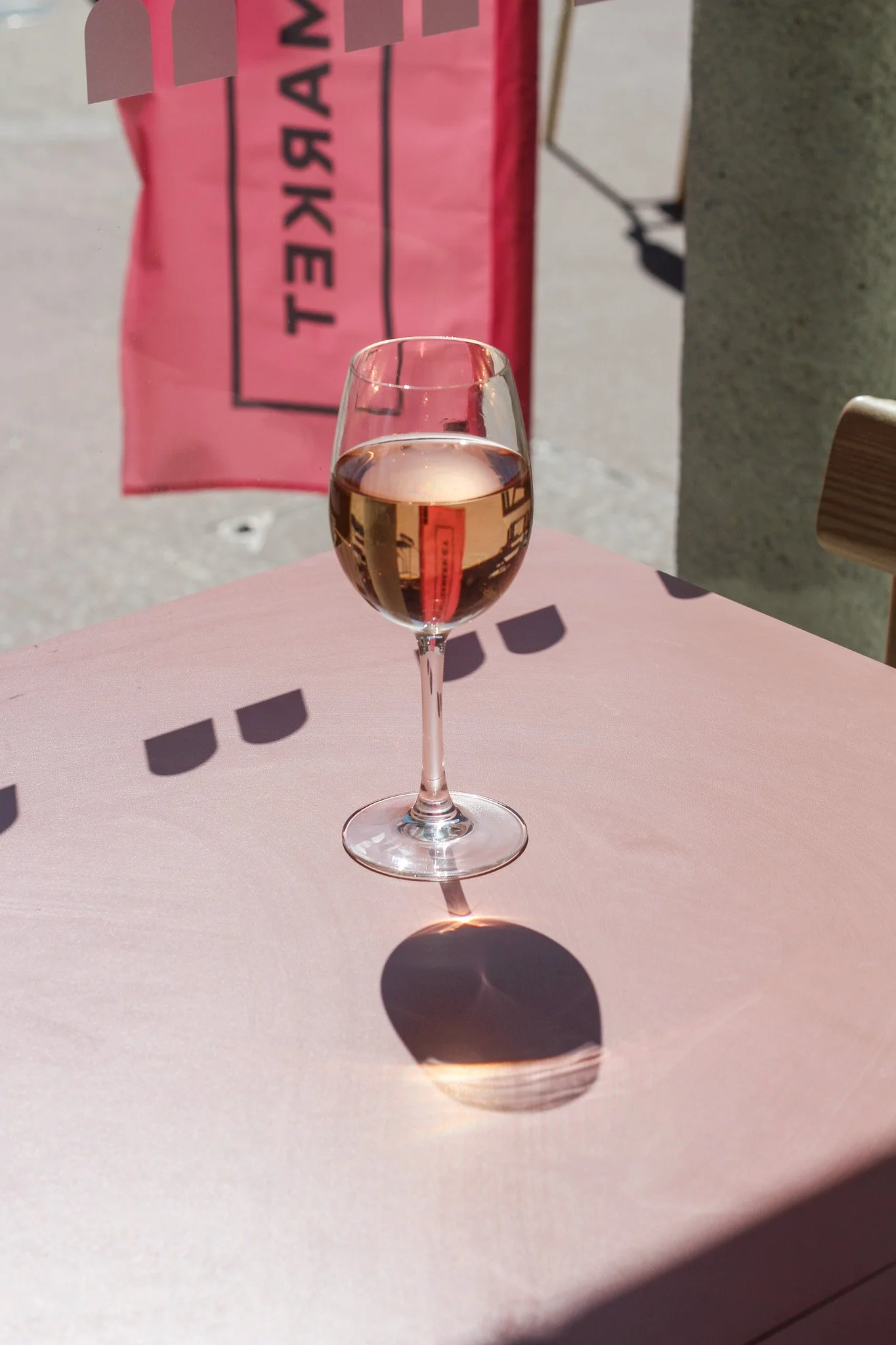 A wine glass filled with pinkish liquid on a pink table outside, with a bright shadow cast by the glass, and pink and gray elements in the background.