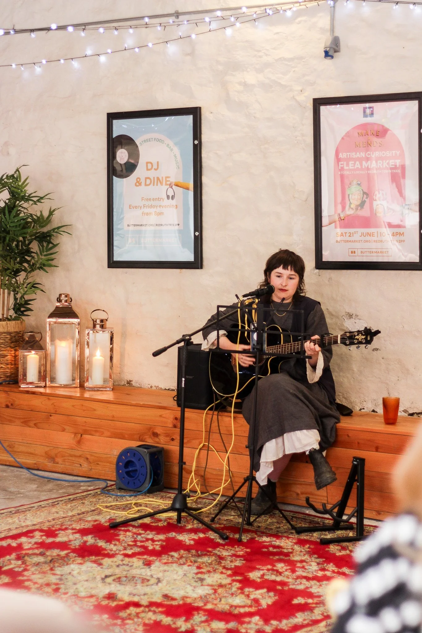 A woman plays guitar and sings into a microphone on a small stage, with a brick wall and posters behind her. There are candles in glass lanterns and a plant on the stage as decoration. The setting appears to be a cozy, intimate live music venue.
