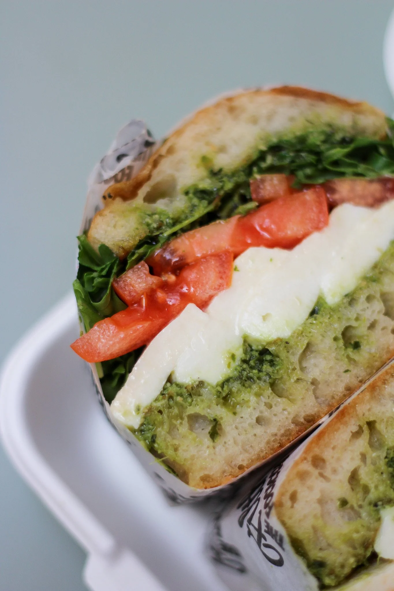 Close-up of a sandwich with layers of tomatoes, greens, cheese, and green sauce, wrapped in paper, in a white container.