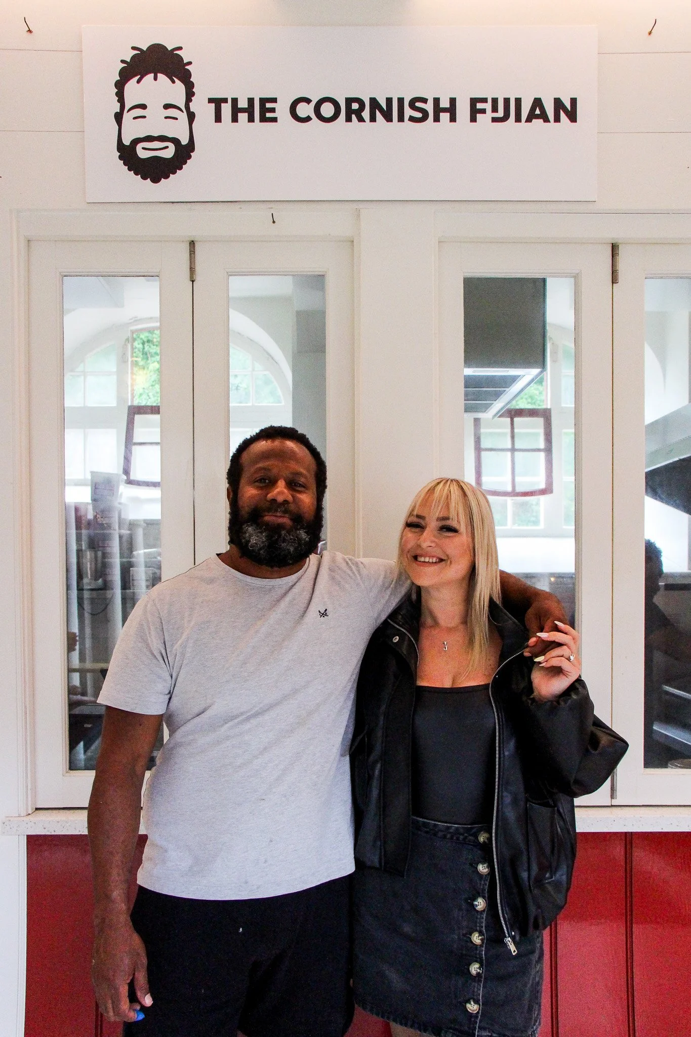 Two people standing side by side in front of a door with a sign above that reads "The Cornish Fijian." The person on the left is a man with dark skin, short curly hair, and a full beard, wearing a light gray t-shirt. The person on the right is a woman with light skin, blonde hair with bangs, wearing a black leather jacket and smiling.